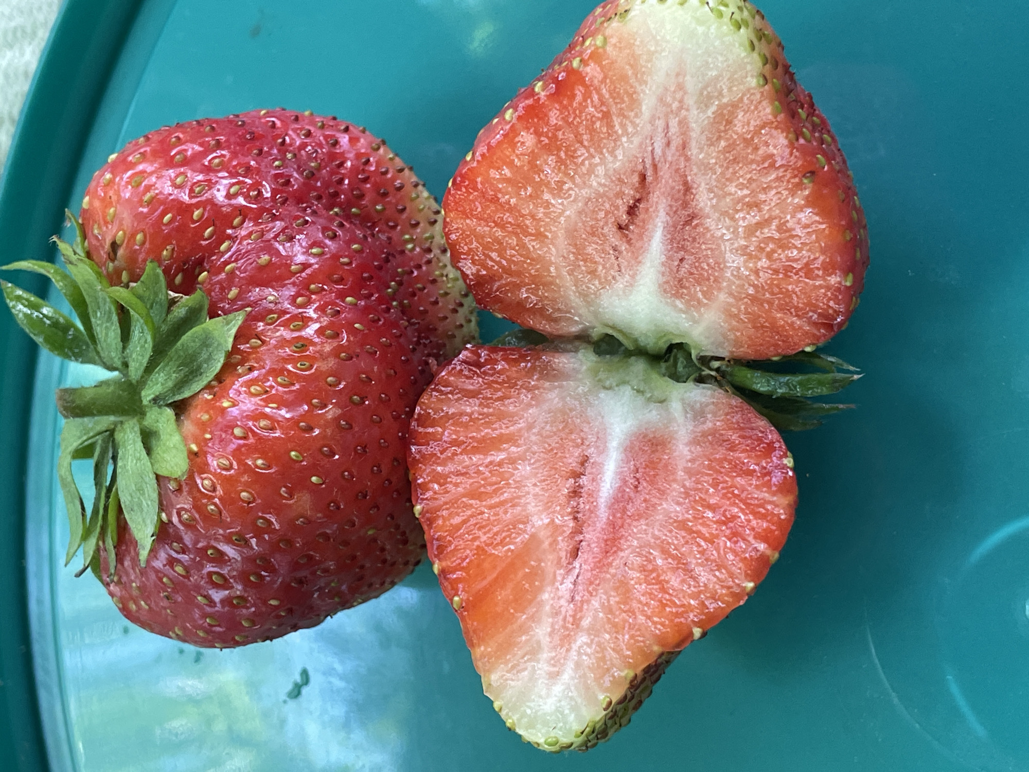 A whole, ripe strawberry with green leaves on top rests on a turquoise platter. To its left, the strawberry is cut in half, revealing its white and pink flesh. The platter is textured with subtle concentric circles. The lighting suggests an outdoor setting, possibly a patio or garden, given the slight hint of an outdoor light source. The overall scene is vibrant and fresh.