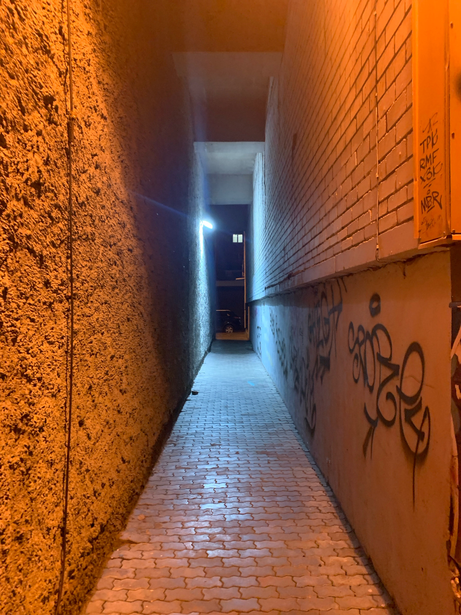 An outdoor, narrow alleyway in Arkhangelsk, Russia, is illuminated by artificial lighting, casting a warm orange glow on the textured wall to the left and a cooler, blueish light on the brick wall to the right. The ground is paved with interlocking bricks, wet from recent rain, reflecting the light. Graffiti adorns the brick wall on the right. At the end of the alley, a dimly lit opening reveals a building interior and a parked car.
