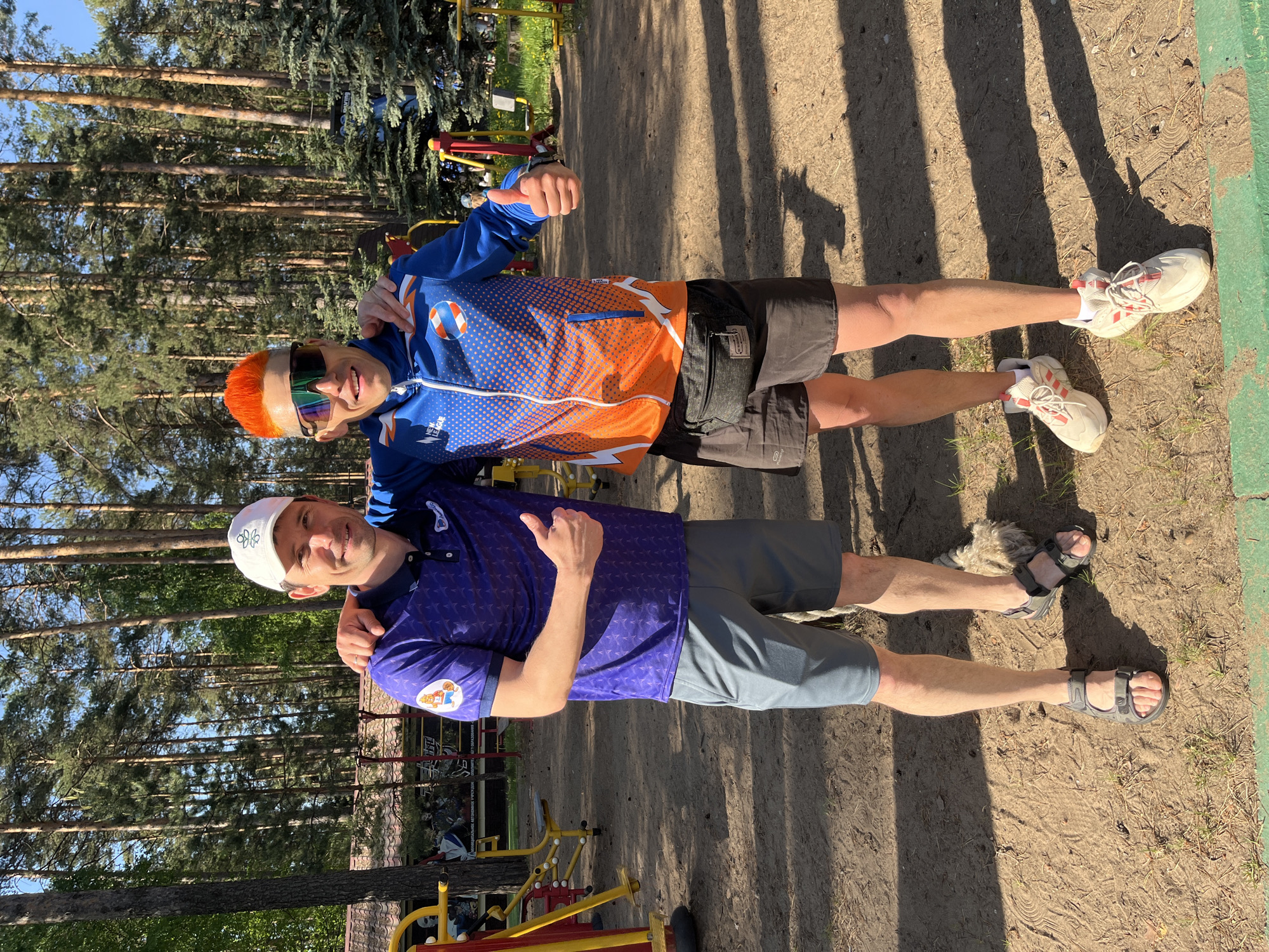 Two men pose for a photo in an outdoor setting. The man in the foreground wears a white baseball cap and a purple collared shirt with a team logo on the sleeve. He wears gray shorts and gray sandals, and he is giving a thumbs-up gesture. The man behind him has orange hair, colored sunglasses, a blue and orange zip-up jacket, black shorts, and white sneakers. He is also giving a thumbs-up. The background shows trees, exercise equipment, and dirt. The location is identified as Chaykovsky, Russia.