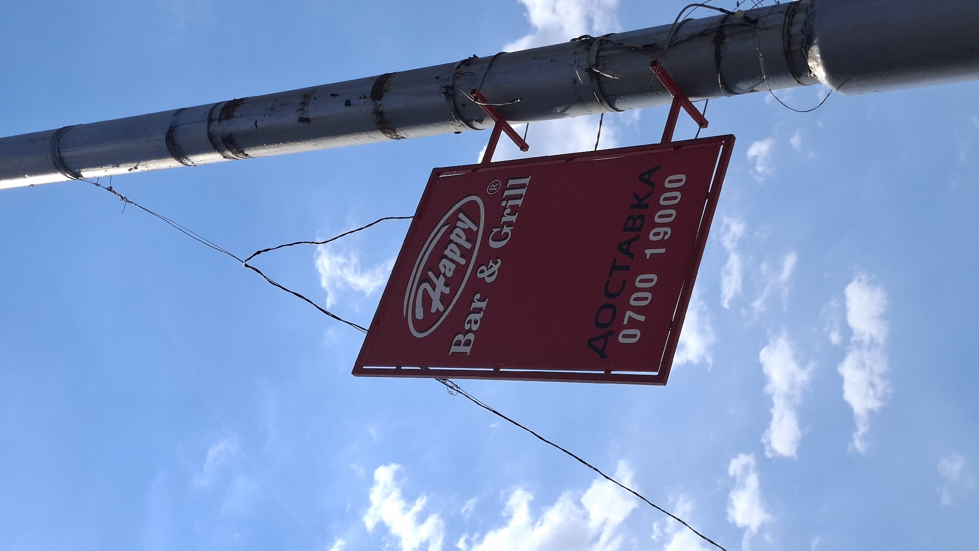 A red sign hangs from a metal pole against a bright blue sky with scattered white clouds. The sign has a white oval logo with the word "Happy" in cursive above the words "Bar & Grill". Below this, in white Cyrillic text, is "ДОСТАВКА" followed by the numbers "0700 19000". The sign is attached to the pole by wires. The pole appears to be a street light pole or utility pole and shows signs of rust and wear. The image is taken from a low angle, looking upwards. The location is not specified in the image.