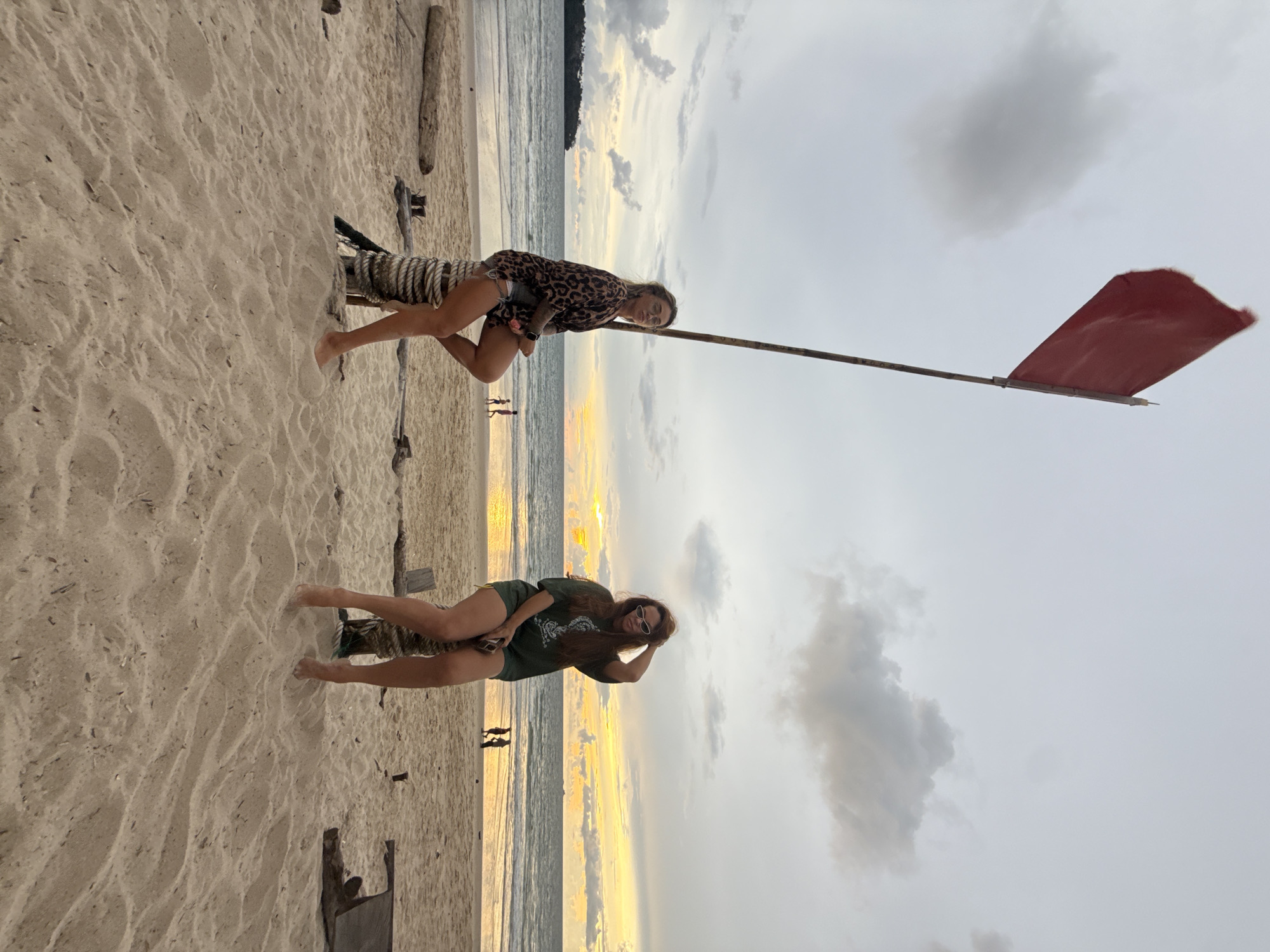 Two women are sitting on rope-wrapped wooden posts on a sandy beach in Choeng Thale, Thailand. A red flag is flying on a tall pole behind them, and the ocean is visible in the background with a sunset casting an orange glow on the water and clouds. The woman on the left is wearing a leopard-print top and shorts, while the woman on the right is wearing a green t-shirt and shorts. Both women have sunglasses on. In the distance, there are a few small figures of people walking on the beach. The lighting suggests it is either early morning or late evening.