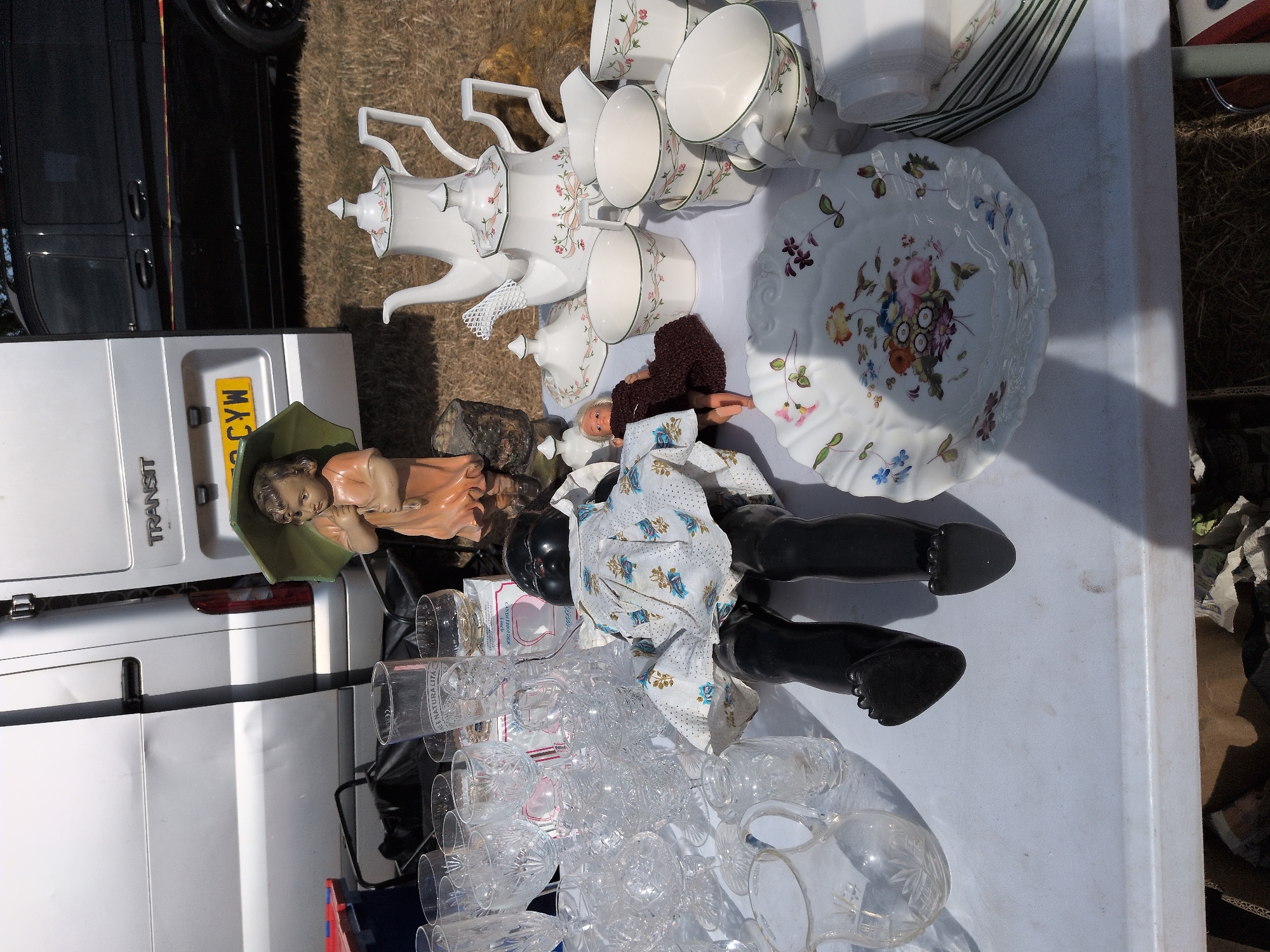 A daytime, eye-level, medium shot captures a white table at an outdoor market in Claverley, United Kingdom. Various items are displayed on the table, including glassware, a porcelain tea set, a doll, and a decorative plate. In the background, a white van and a black van are visible, parked on a dry, grassy surface. The scene is well-lit, with shadows suggesting the sun is positioned to the right of the frame.