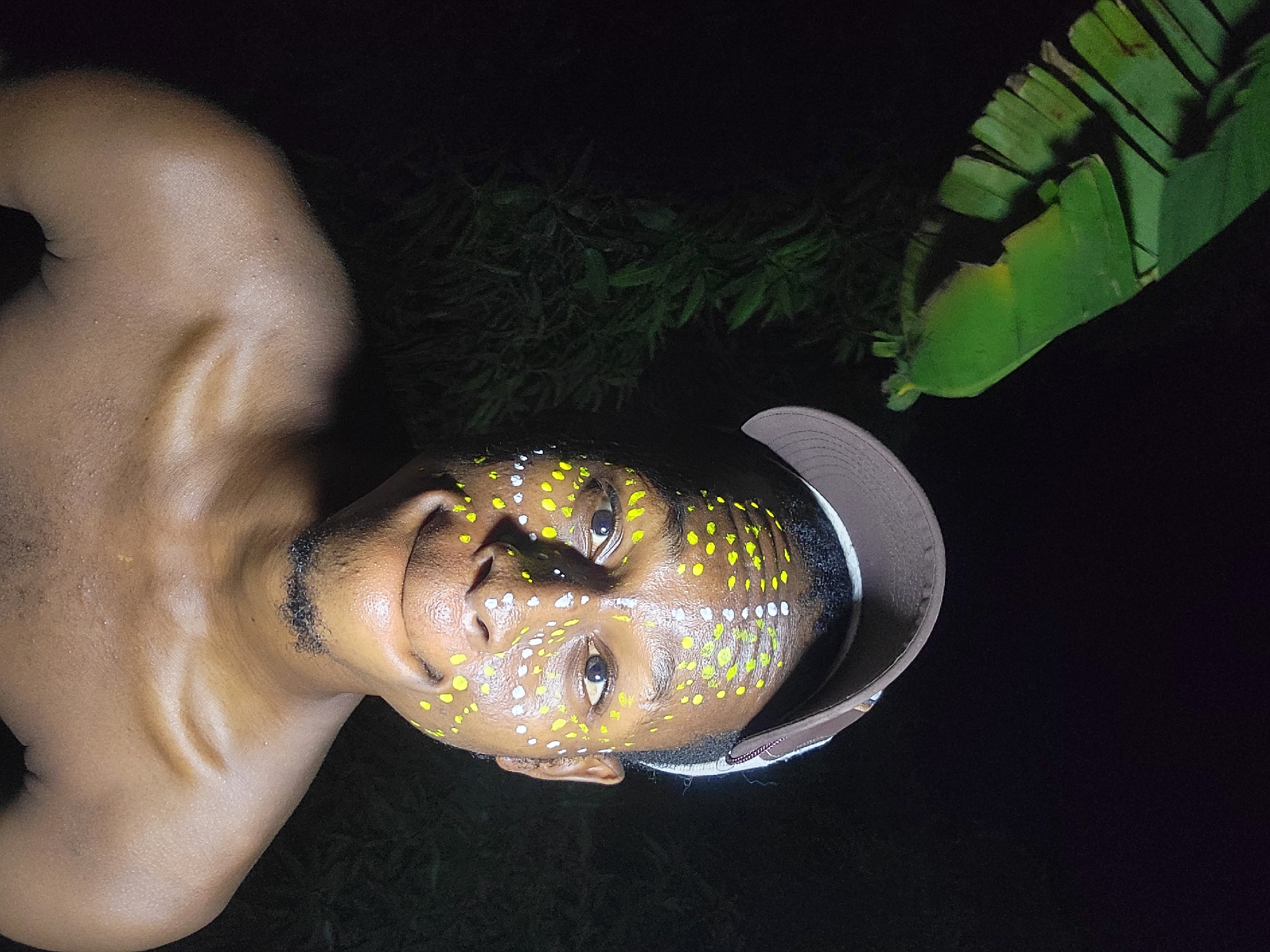 A shirtless man with yellow and white dots painted on his face and wearing a baseball cap is photographed at night in Nkpor, Nigeria. The background is dark with some green foliage including a large leaf visible in the upper right corner. The man's face is lit and he is looking directly at the camera with a slight smile.