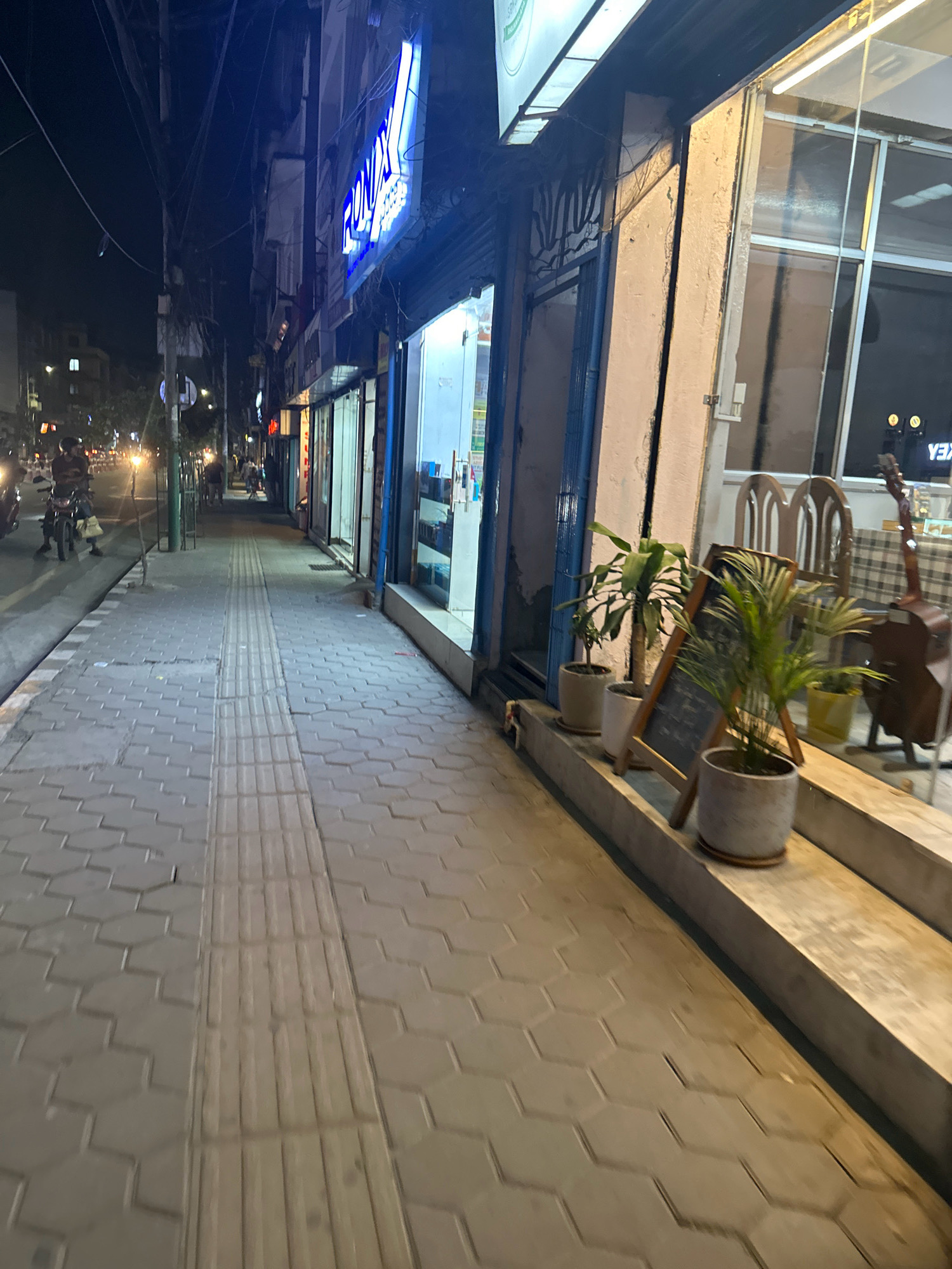 The image depicts a street scene at night in Kathmandu Metropolitan City, Nepal. A sidewalk paved with hexagonal tiles and a tactile paving strip runs along the right side of the frame. Several potted plants are displayed outside a shop, accompanied by a chalkboard sign. Further down the sidewalk, there are chairs and tables, and a guitar leaning against a wall. The street is illuminated by streetlights and the lights from the shops, and there is some traffic visible in the distance, including motorcycles. A prominent blue neon sign with white lettering that reads "RONIX" is visible on the facade of one of the buildings. The overall environment appears to be an urban commercial area.