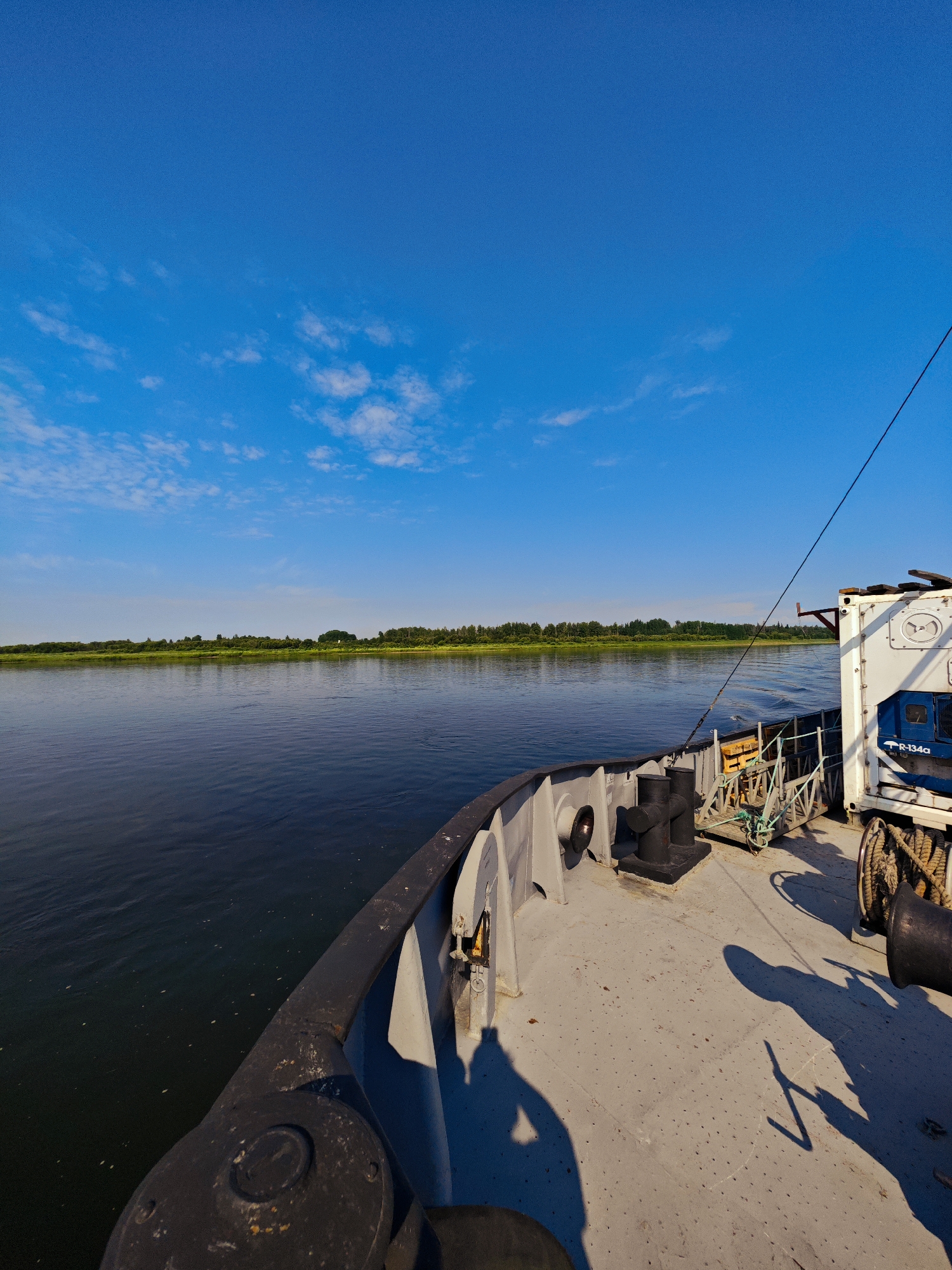 This image depicts a view from the deck of a vessel on a river, likely in or near Novonazimovo, Russia, given the context. The sky is a clear, vibrant blue with sparse, wispy white clouds. To the left of the frame, a calm, dark river stretches towards a distant, tree-lined shore. The shore is a continuous band of green foliage, suggesting a natural landscape.
The foreground is dominated by the deck of the vessel, which is made of a gray, weathered material. Several objects are visible on the deck, including large black bollards, a coiled rope on a spool, and a white shipping container with blue accents and visible lettering that reads "R-134a". Shadows of what appear to be people are cast across the deck, indicating the presence of at least two individuals. One shadow is particularly distinct, showing a figure with an arm raised. A black railing runs along the edge of the deck. The vessel appears to be in motion, with subtle ripples visible on the water's surface. The overall atmosphere is serene and suggests a peaceful journey on the water under a bright sky.
