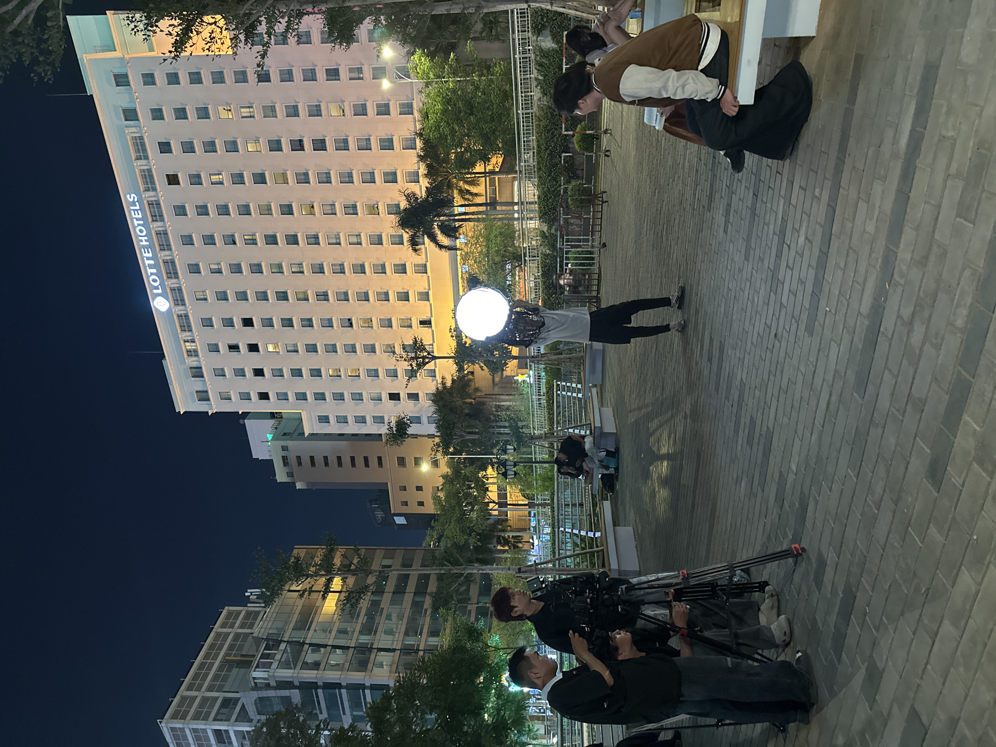 The image shows an outdoor evening scene in Thủ Đức, Vietnam, featuring a film crew working in front of the Lotte Hotel. A tall, white building with many illuminated windows is visible, bearing the "LOTTE HOTELS" logo. The film crew consists of several individuals, including two men operating a camera on a tripod and a person holding a large, round light source. Another person, possibly an actor or director, is kneeling and appears to be reviewing a script or scene. The setting includes paved walkways, trees, and other buildings in the background. The lighting is a mix of artificial lighting from the film equipment and streetlights, creating a brightly lit focal area amidst a darkening environment.