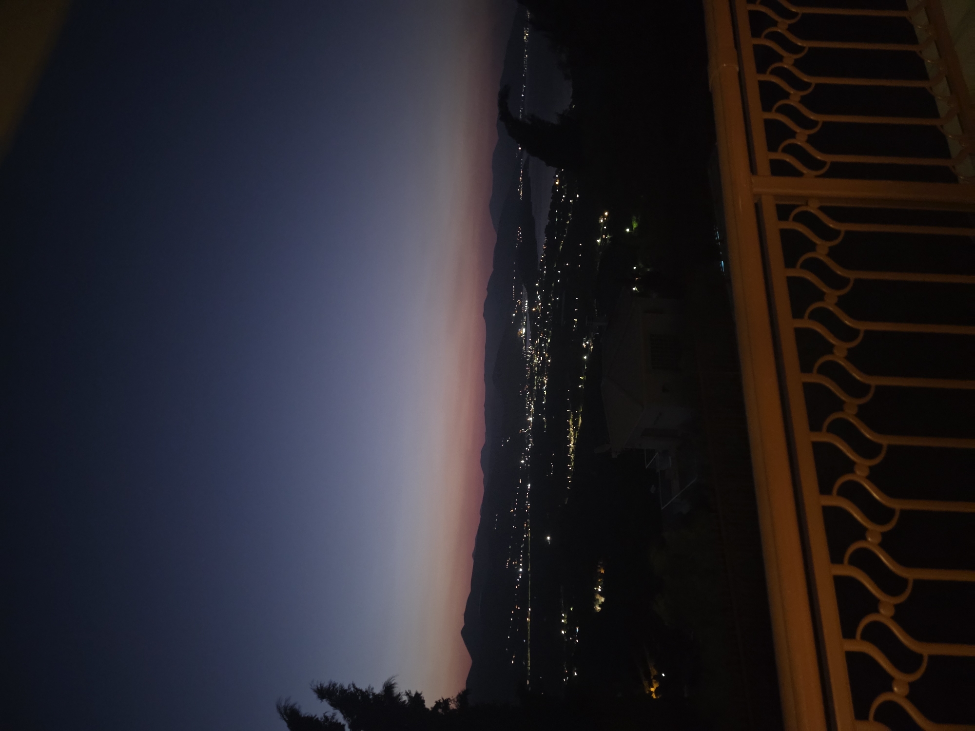 A vertical, outdoor, low-light, telephoto shot captures a twilight view over the Municipal Unit of Asini, Greece. The sky transitions from deep indigo at the top to a faint pink and orange hue near the horizon, where a silhouetted mountain range dominates the background. Below the mountains, a town is illuminated by numerous small, bright lights, suggesting a coastal settlement with a body of water visible to the right. In the foreground, a decorative, gold-colored metal railing with ornate scrollwork partially obscures the lower portion of the scene. Dark, silhouetted trees frame the left and right sides of the image, with a hint of a building visible on the far right. The overall impression is one of a serene, twilight landscape.