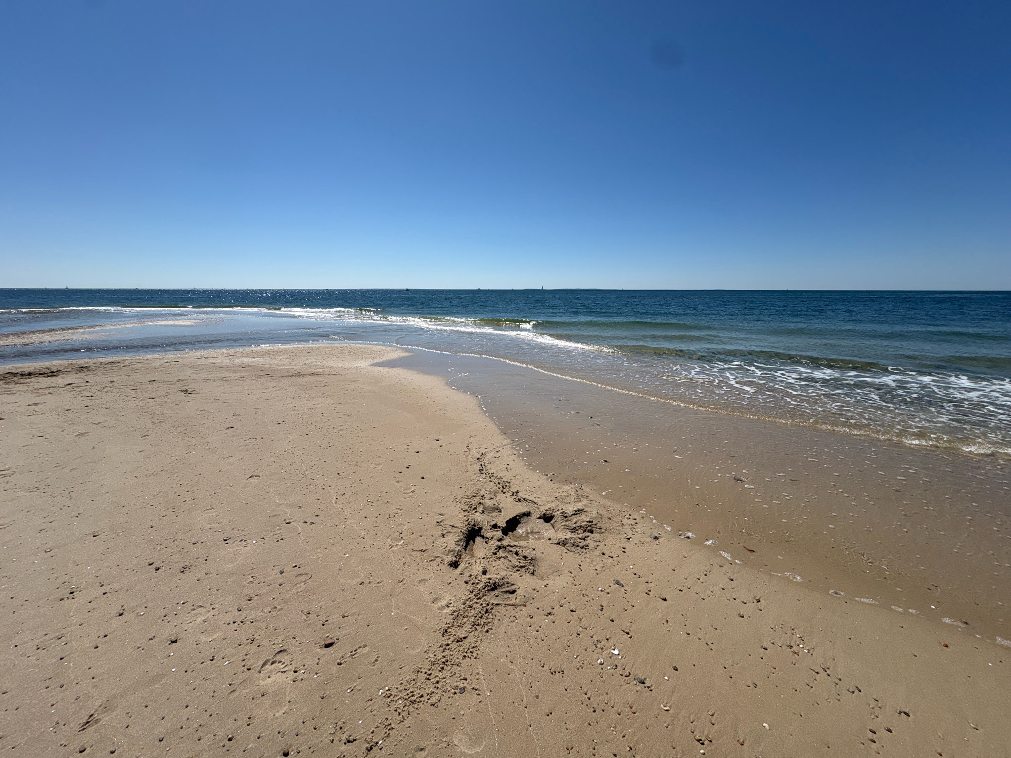 The image shows a sandy beach with the ocean in the background. The sky is clear and blue. There are some footprints in the sand. The scene is peaceful and serene. This is a generic beach scene and cannot be identified as being in Waterford, United States of America.