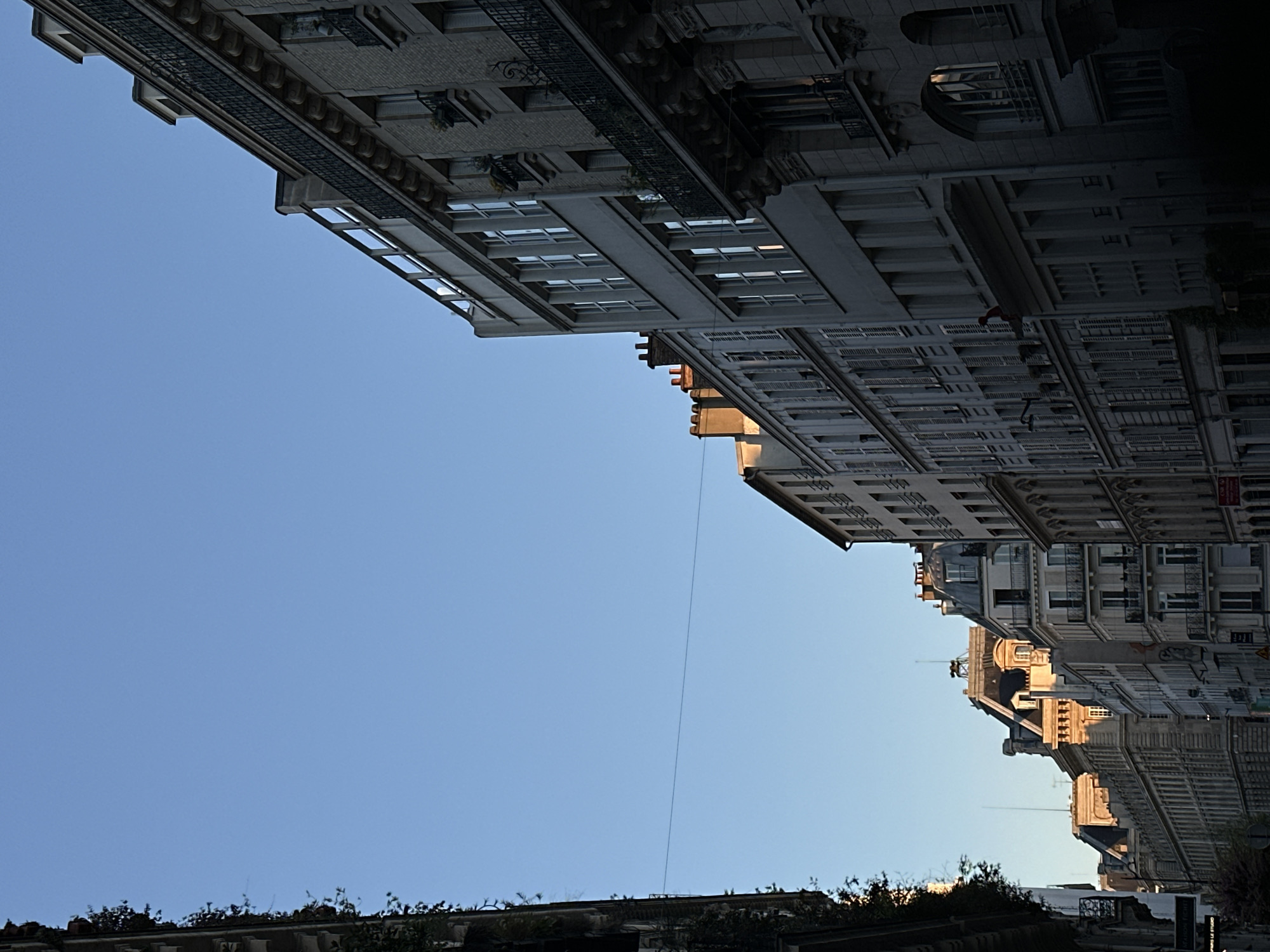 The image shows a street in Paris, France, with tall buildings lining both sides. The buildings have intricate architectural details, including rows of windows, balconies, and ornate facades. The sky is a clear blue, with a gradient of light from the horizon. Trees and greenery are visible at the bottom of the image, along the street level. A thin wire or cable stretches across the sky between the buildings.