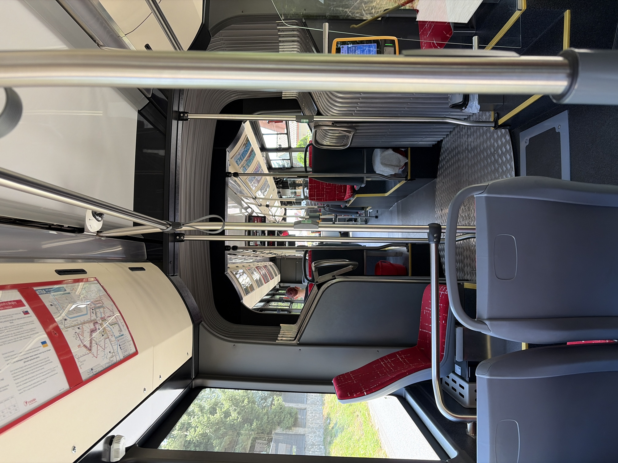 The image shows the interior of a public transport bus. The bus appears to be modern, with grey plastic seats and red fabric seats with a patterned design. There are metal poles throughout the bus for passengers to hold onto. A map of bus routes is displayed on the ceiling at the front of the bus. Sunlight streams in through the large windows, illuminating the interior. Outside the windows, trees and buildings are visible, suggesting an urban or suburban environment. A person can be seen sitting at the back of the bus, facing away from the camera. The flooring is dark with a textured metal section in the aisle. A ticket machine is visible on the right side of the bus. Based on the map and signage in the bus, it is likely operating within the city of Nitra, Slovakia, as indicated by the word "NITRA" visible on the route map.