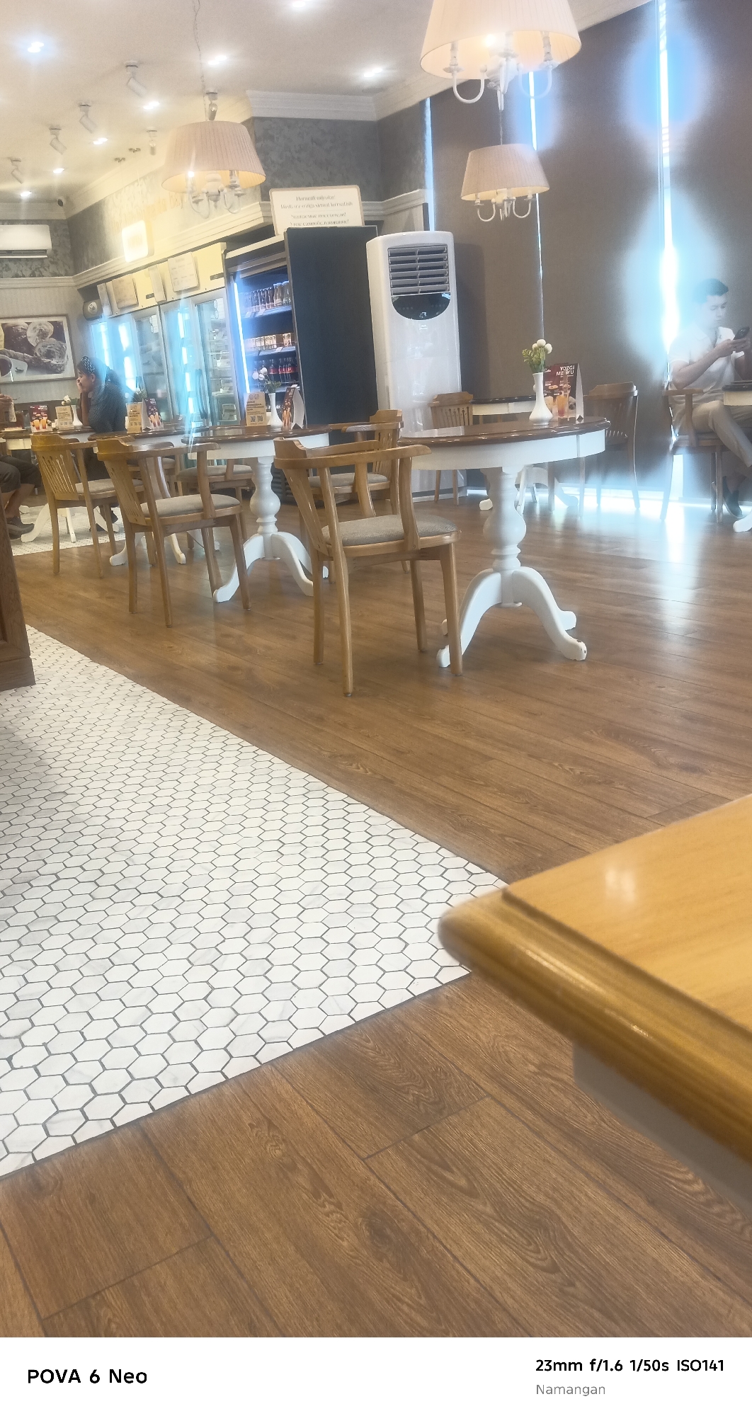 The image depicts the interior of a well-lit cafe or restaurant in Namangan City, Uzbekistan. In the foreground, a polished wooden floor transitions to a white hexagonal tile floor. Several round tables with white bases and wooden chairs are arranged throughout the space. In the mid-ground, a tall white air conditioning unit stands near a refrigerated display case filled with food items. A woman is seated at a table on the left, looking down. On the right side of the image, a man is seated at a table, looking at his phone. The background shows more tables and chairs, with soft lighting emanating from ceiling fixtures and a bright window or glass door. The overall ambiance suggests a clean and modern dining establishment.