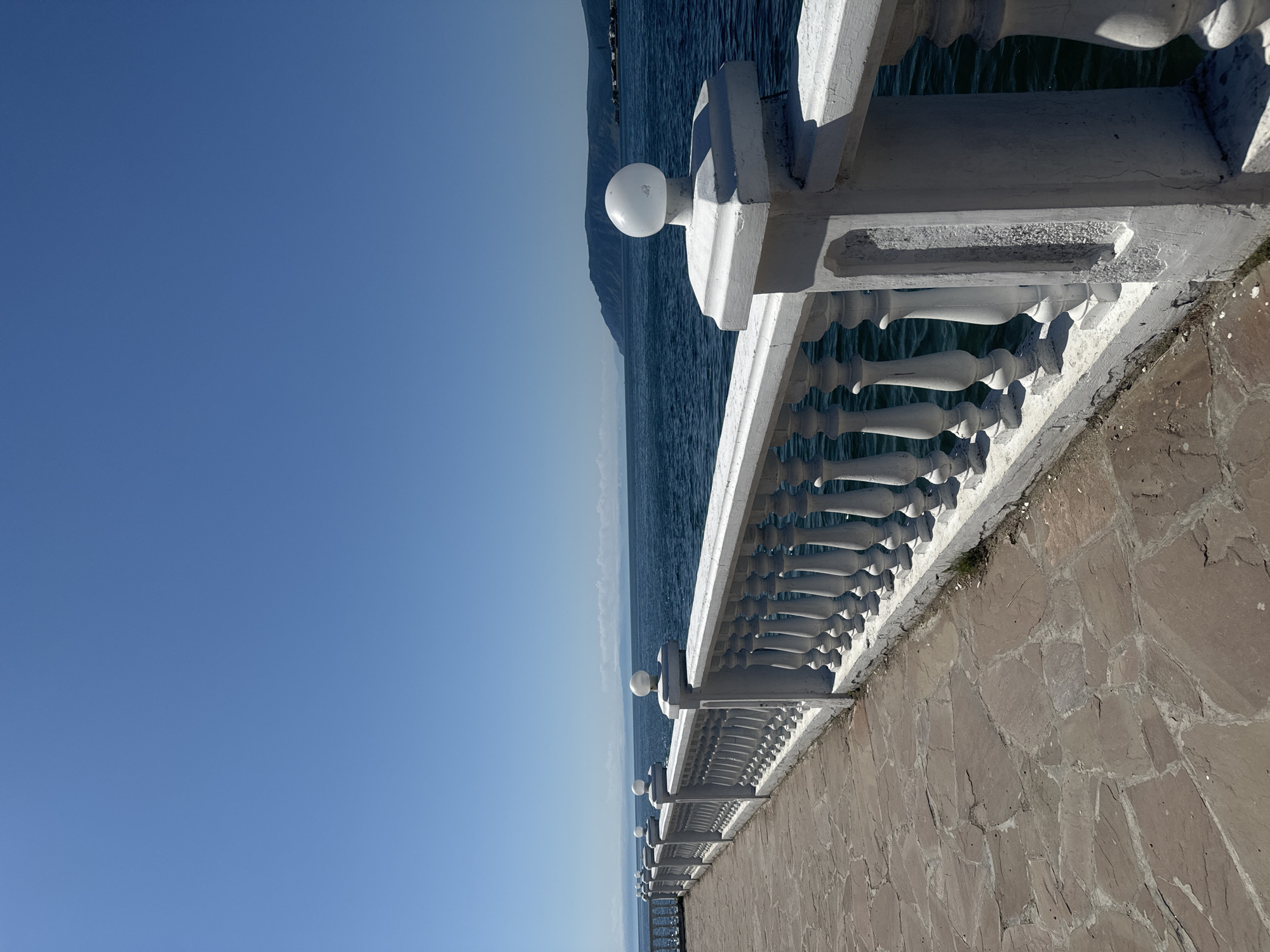 This is a vertical, eye-level, medium shot of a white, ornate balustrade bordering a stone-paved walkway alongside a large body of water. The balustrade is made of individual balusters and topped with a handrail. White, spherical light fixtures are mounted on decorative posts along the balustrade. The walkway is paved with irregular, reddish-brown flagstones. Beyond the balustrade, the dark blue water stretches to the horizon, where a distant, hazy shoreline with low hills is visible under a clear, bright blue sky. The location is identified as the Listvyanka embankment in Listvyanka, Russia. There are no people or animals visible in the image. The scene depicts a tranquil, sunny day with a clear view of the water and distant landscape.
