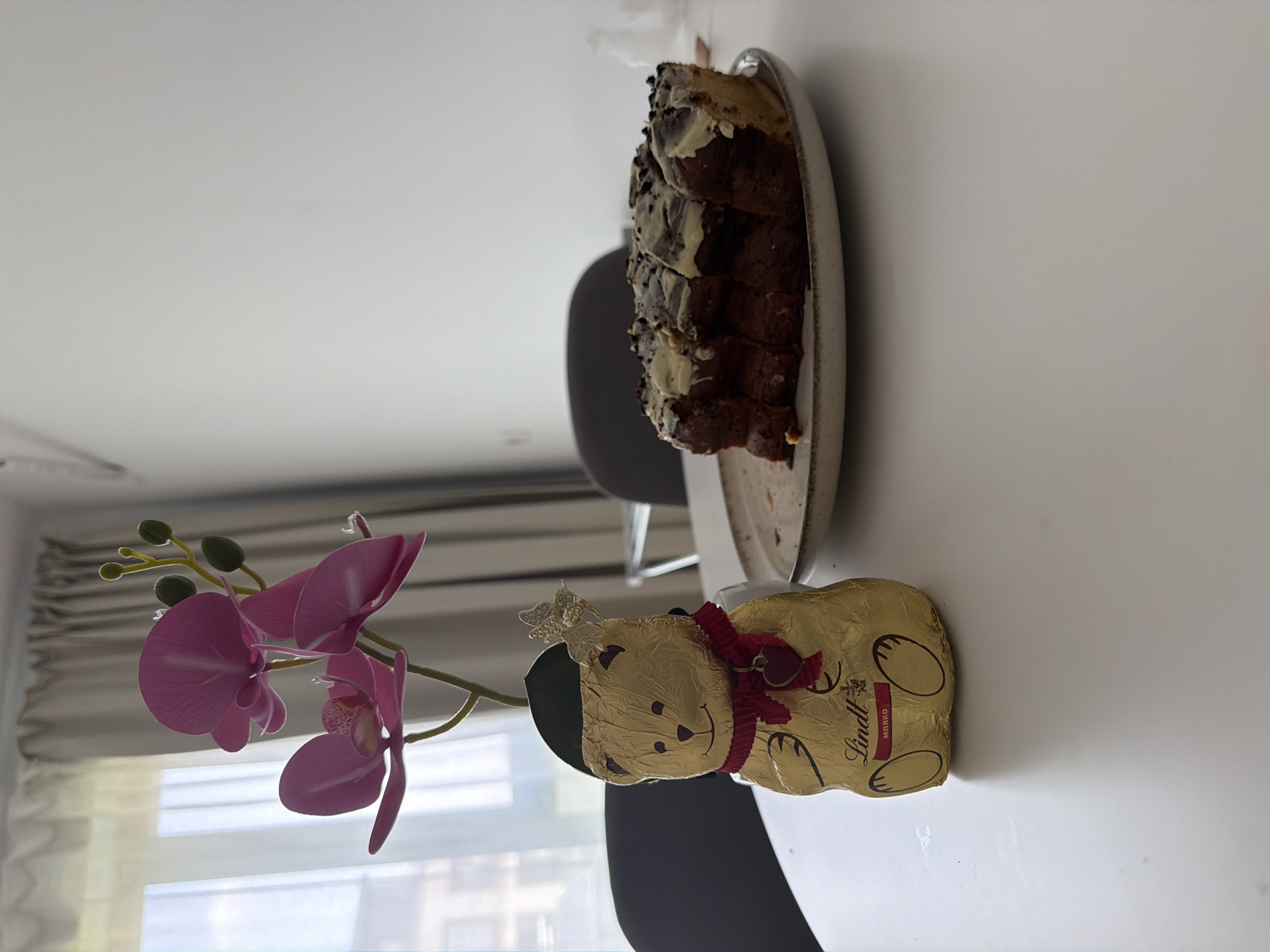 The image depicts an indoor scene within a residential apartment, likely situated in Sofia, Bulgaria. The primary focal point is a white, rectangular table serving as the central surface.
On the left side of the table, a gold-foil-wrapped Lindt chocolate bear stands upright. The bear is adorned with a red fabric scarf featuring a small, red heart-shaped charm. A decorative gold butterfly clip is attached to the bear's head, positioned in front of a dark, hat-like embellishment. The Lindt logo and the Cyrillic text "МЯРКО" are visibly printed on the bear's foil, the latter strongly indicating a Bulgarian market presence due to its similarity to "мляко" (milk). Behind the chocolate bear, an artificial pink orchid plant with multiple blooms and green buds emerges from a dark, unseen pot, arranged so it appears to be growing from behind the bear.
On the right side of the table, a ceramic plate holds a partially consumed section of a dark brown cake or dessert. The cake features several distinct layers or slices, topped with irregular streaks of white icing and dark sprinkles or crumbs. A noticeable portion of the cake is missing, indicating it has been eaten. The plate is light-colored with a rustic, textured rim.
The background on the left reveals a large window covered by sheer white curtains, allowing diffused natural light to illuminate the scene. Beyond the curtains, a blurry urban environment with buildings is faintly visible. The background on the right consists of a plain white wall. A subtle fixture, possibly an air conditioning unit or smoke detector, is partially visible high on this wall. Two dark-upholstered chairs are partially visible in the midground, suggesting a dining or seating area. No individuals are present in the image, and no actions or interactions are depicted. The overall lighting is bright and even, consistent with natural daylight.