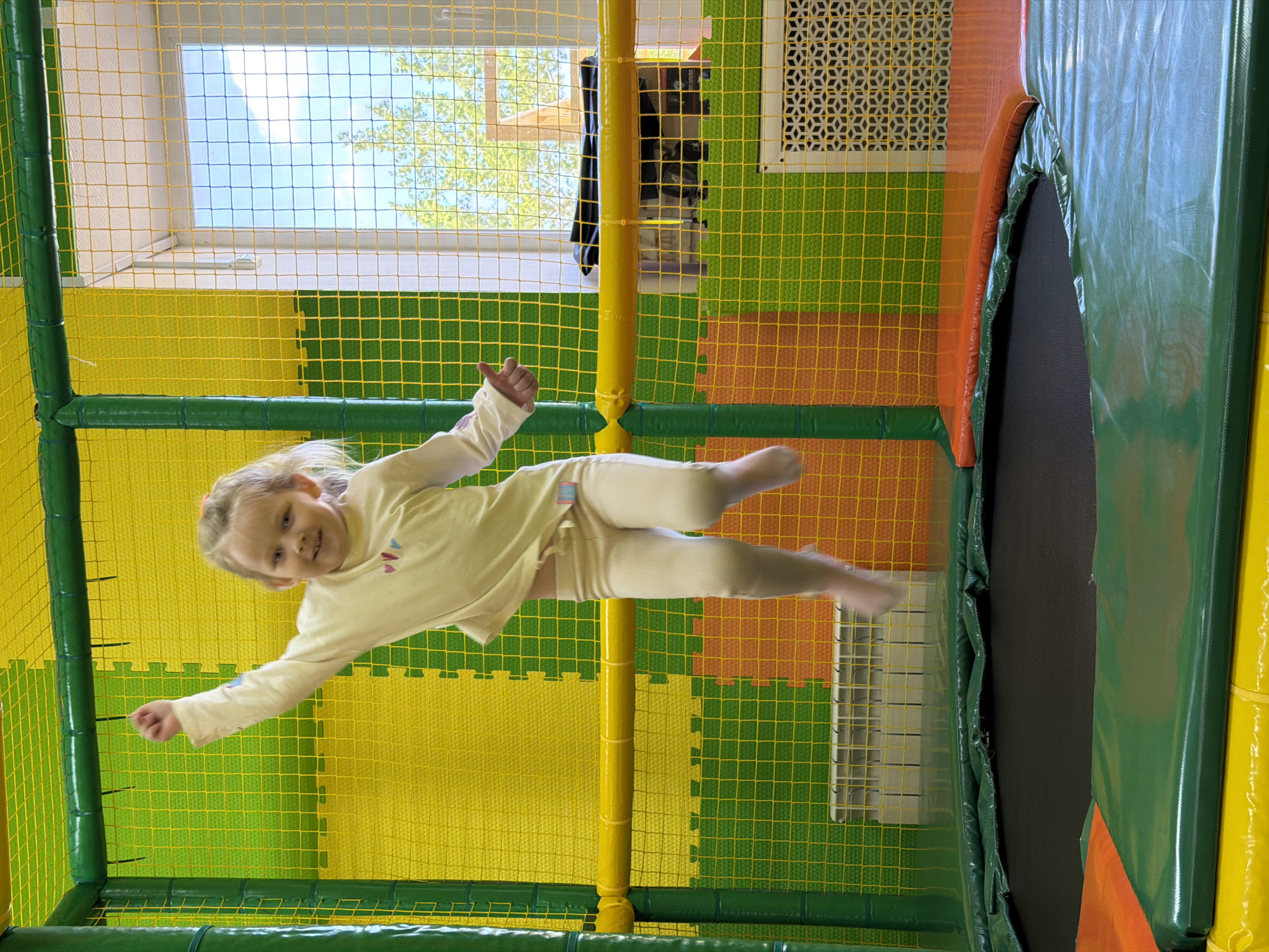 A young girl is captured mid-air in an indoor playground. She is wearing a light-colored top and pants and has her arms extended outwards. She appears to be jumping on a trampoline. The playground has colorful yellow and green padded floors and walls covered in a net. In the background, there is a window with a view of trees and the sky. The image was taken in Kamyshin, Russia.