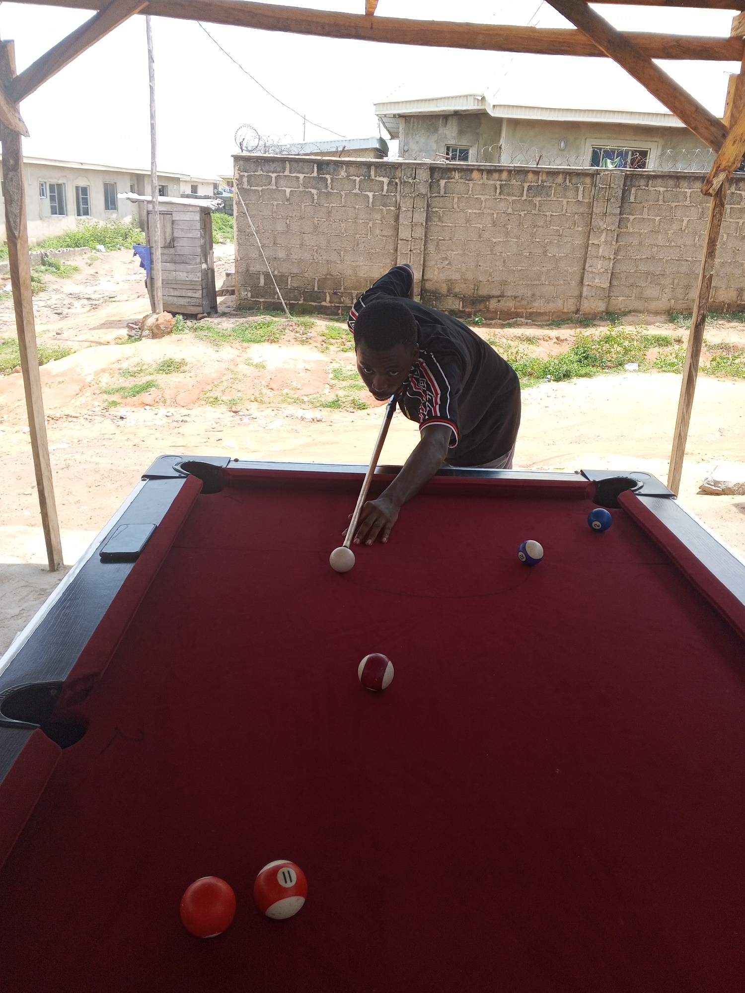 A young Black male is playing pool outdoors in Lokoja, Nigeria. He is wearing a black t-shirt with red and white stripes on the sleeves and is bent over the pool table, holding a cue stick and aiming at a white cue ball. The pool table has a red felt surface and black trim, with several balls scattered across it, including a ball with the number 11. The background shows a brick wall topped with barbed wire, several residential buildings, and a clear sky. The area appears to be an unpaved, dusty lot.