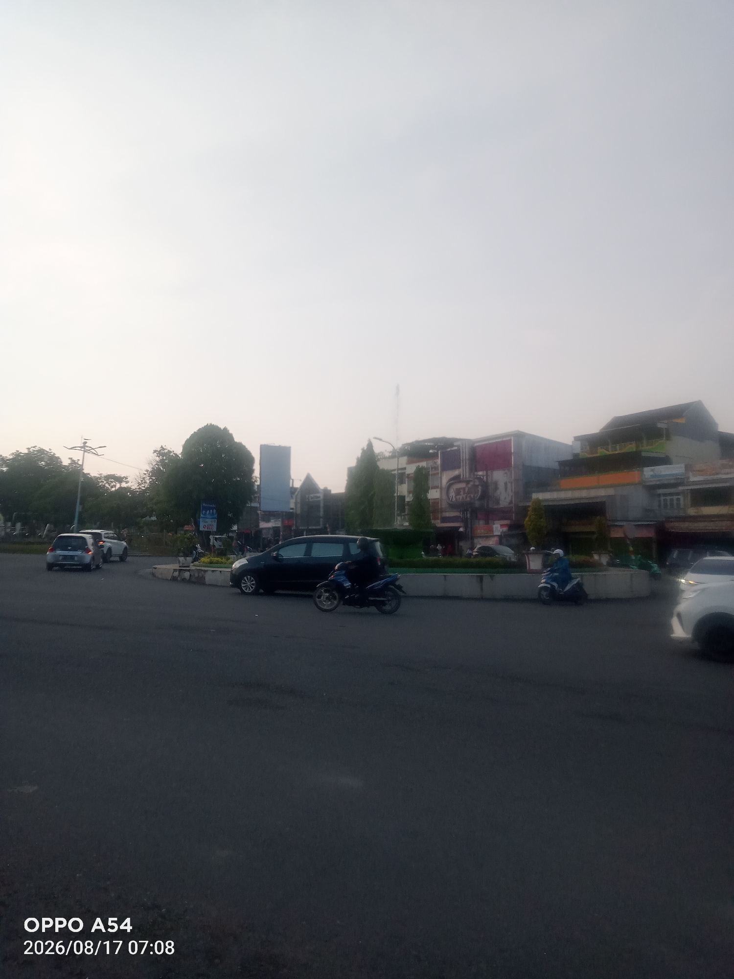 The image depicts a street scene in Jambi, Indonesia, likely at dawn or dusk, given the soft, overcast lighting. The foreground shows a paved road with a central divider. Several vehicles are present: a dark-colored sedan is turning right, a motorcycle with a rider is proceeding forward, and another motorcycle with a rider is on the right side of the road. In the background, buildings line the street, including a multi-story structure with signage that appears to be a cinema or entertainment venue, and other commercial establishments. Trees are visible, adding greenery to the urban landscape. The sky is a uniform pale gray. A timestamp in the bottom left corner indicates the image was captured on "2026/08/17 07:08" by an "OPPO A54" device.