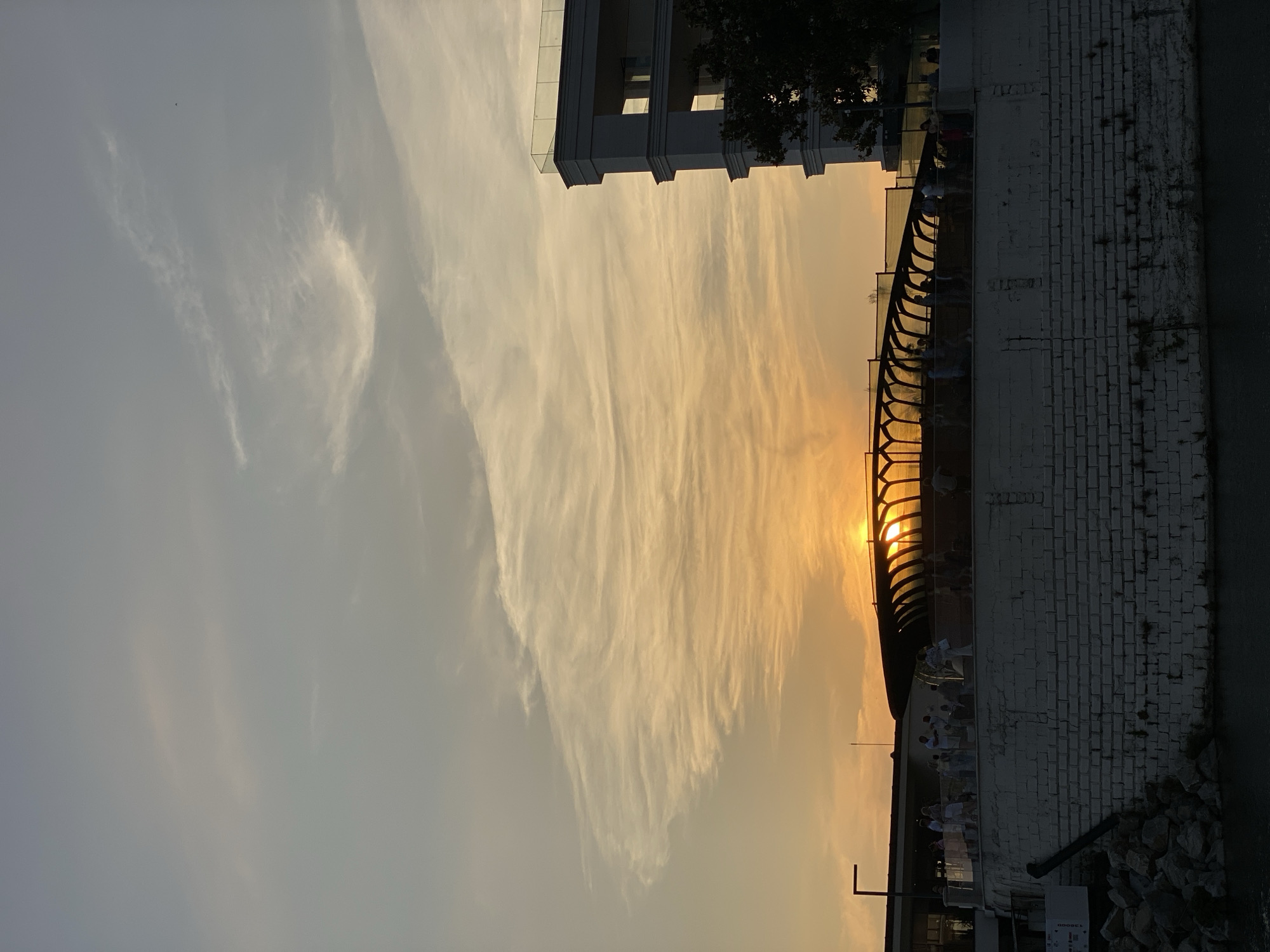 This is a photograph of a sunset at the Sochi Sea Port in Sochi, Russia. The sun is low in the sky, casting a warm golden glow on the clouds and the surrounding buildings. In the foreground, there is a white brick wall with a body of water in front of it. On top of the wall, a group of people are gathered, walking along a promenade that is covered by a modern, arched structure with glass panels. To the right, a white, multi-story building with balconies is visible, and a dark tree is in front of it. The sky is filled with wispy clouds, painted with streaks of orange and yellow from the setting sun.