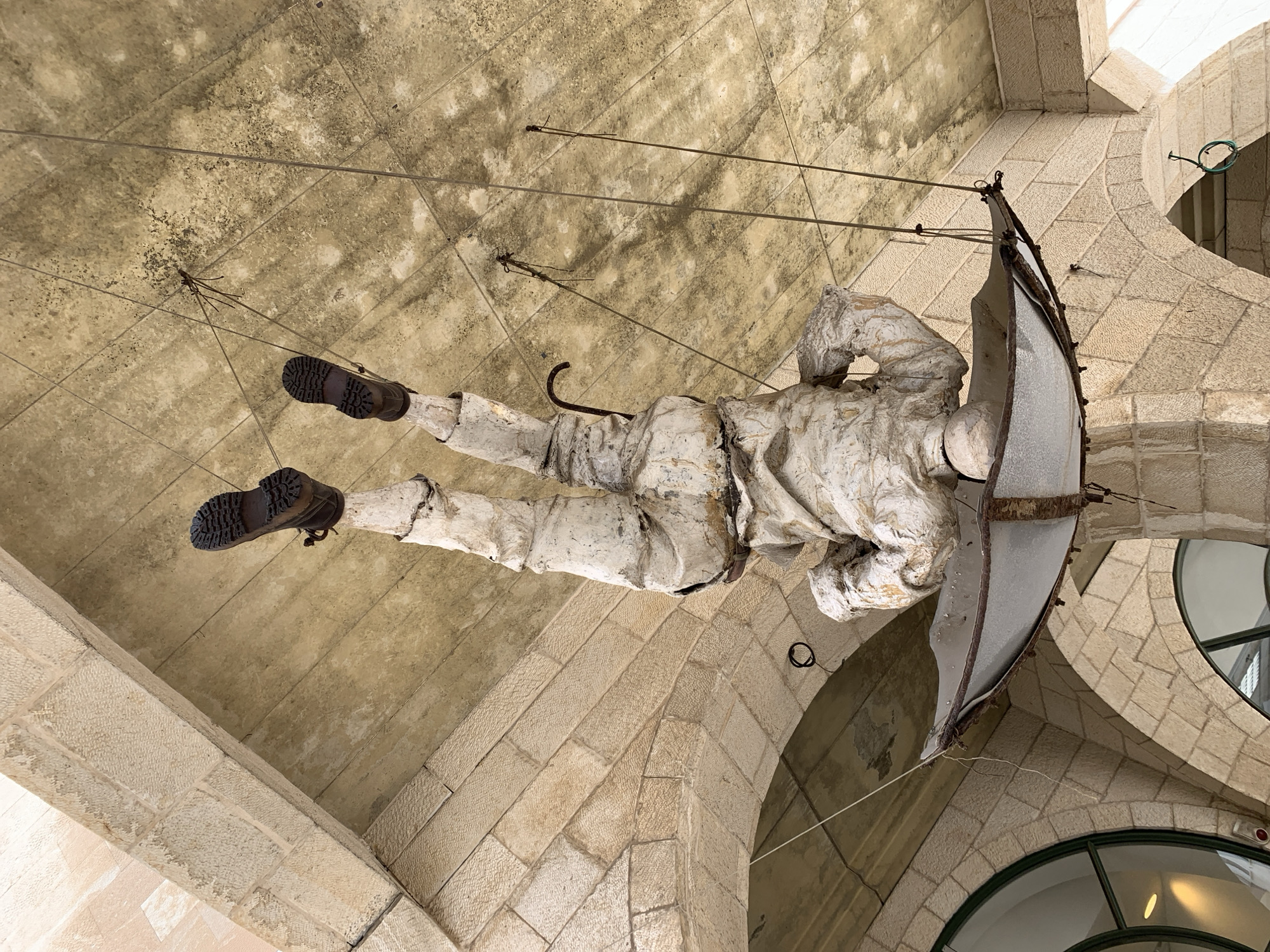 An art installation depicting an upside-down humanoid sculpture is suspended from the ceiling within an arched stone structure. The sculpture, rendered in a light-colored, textured material resembling wrapped fabric or plaster, shows a figure with legs extended upwards, wearing dark brown, lug-soled boots. Its torso is positioned downwards, partially encased within a large, inverted, damaged umbrella-like structure, also light-colored with a dark frame, and a dark, curved handle protrudes from the figure's chest area. Multiple thin wires or rods suspend the entire assembly from a mottled, light-tan, tiled ceiling. The surrounding architectural environment consists of light-colored, roughly cut stone blocks forming multiple arches and support beams, with arched windows featuring dark frames visible in the background. This scene is located in Old Jaffa (Yafo), Tel-Aviv, Israel.