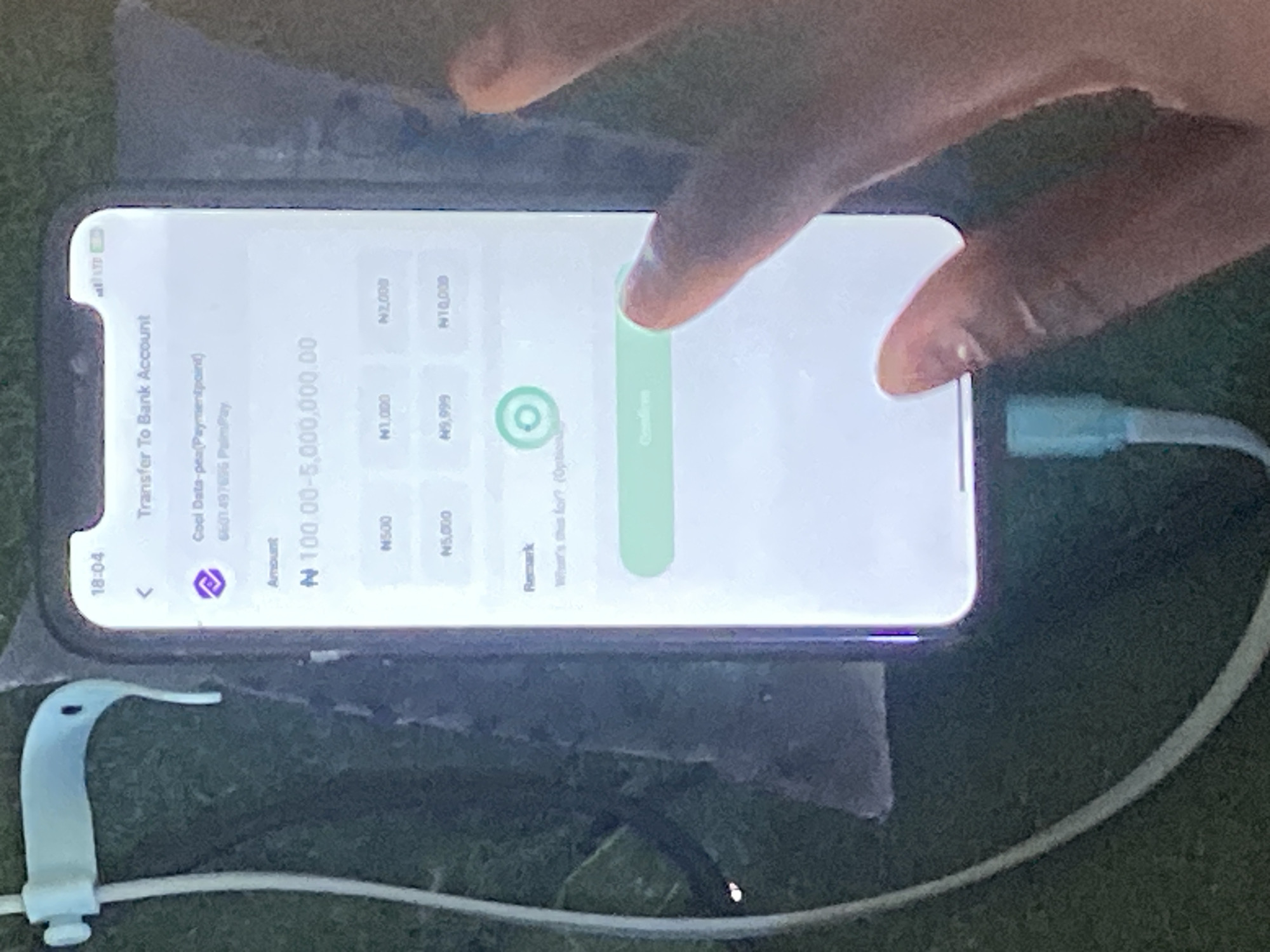 A close-up, slightly blurry photograph captures a person's hand interacting with a smartphone screen displaying a financial transfer interface. The phone is angled and held in the foreground, with a thumb positioned over the "Confirm" button. The screen shows details for a transfer to "Cool Data-pay (Paymentpoint)," with an amount field displaying "N 100.00-5,000,000.00." Several pre-set amount options like N500, N1,000, N2,000, N5,000, N9,999, and N10,000 are visible. A "Remark" section is present with an optional input field. The phone is connected to a blue charging cable. In the background, a green, textured surface, possibly grass or a mat, is visible. The overall lighting suggests an outdoor or well-lit indoor environment. The image provides no specific visual cues to pinpoint its location in Jauro Yinu, Nigeria, other than it being a place where this financial transaction is being conducted via a smartphone.