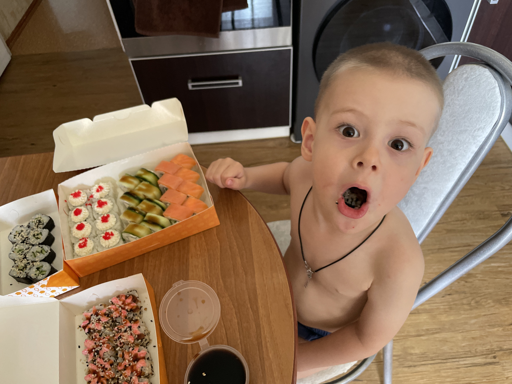 A high-angle shot shows a young, shirtless boy with short brown hair and a necklace eating sushi at a table laden with takeout sushi containers. The boy's mouth is open, revealing the sushi inside. On the wooden table are three white takeout containers with different types of sushi, including rolls with salmon, cucumber, and imitation crab. Two small containers, one with a light-colored sauce and the other with dark soy sauce, are also present. Behind the table are a cabinet with a stainless steel door and a dark-colored appliance. The boy is seated on a silver metal chair with a white seat. The floor is light brown with wood grain.