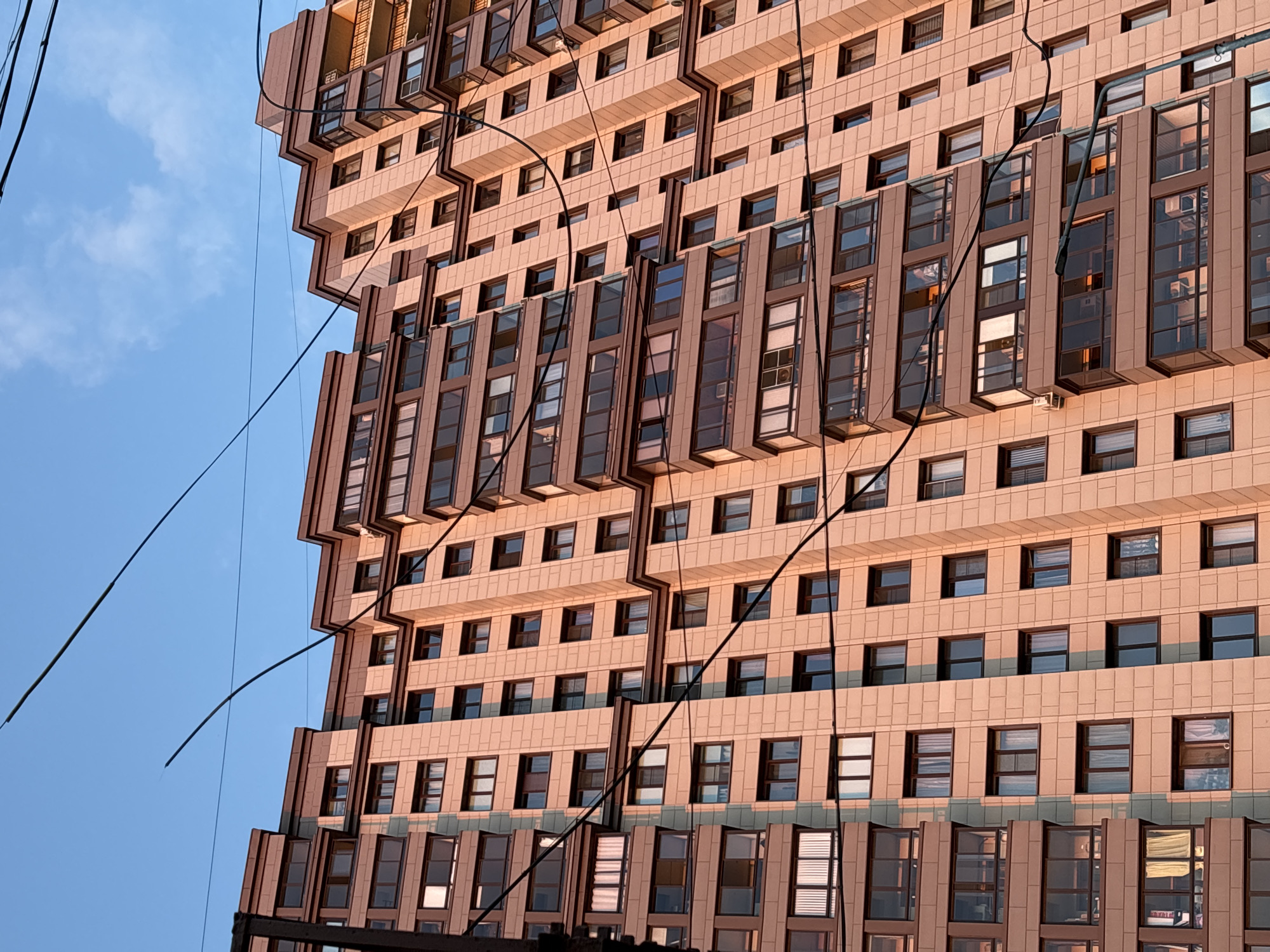 The image captures a view of a modern apartment building in Nizhny Novgorod, Russia, under a clear blue sky with some wispy clouds. The building is tall and multi-story, with a facade predominantly made of light-colored, textured panels that appear to be sandstone or a similar material. The windows are numerous and arranged in a grid pattern, with some featuring dark frames and tinted glass. Several balconies are visible, some enclosed with glass, others open. The building's architecture features stepped upper levels with darker colored accents and protrusions.
Several black electrical wires are strung across the frame, crossing in front of the building at various angles, adding a sense of urban infrastructure to the scene. The lighting suggests it is either early morning or late afternoon, as the sunlight casts a warm, golden hue on the building's facade, creating soft shadows. A street light is partially visible on the right side of the image. No people are clearly identifiable in the image, though some reflections in the windows might suggest interior activity. The perspective is from below, looking up at the imposing structure.