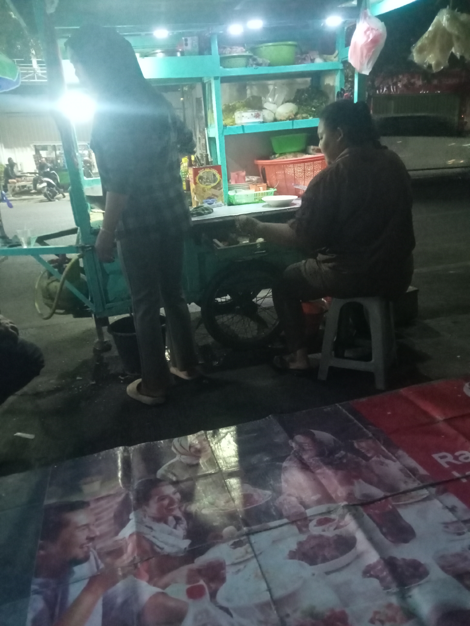 This is a night scene of a street food vendor in Probolinggo, Indonesia. The vendor, a person with dark hair wearing a brown shirt, is sitting on a grey stool behind a teal-colored food cart. The cart is equipped with shelves holding various food items and containers, including what appears to be fresh produce and packaged goods. A metal gas cylinder is visible attached to the cart. In the foreground, a poster is laid out on the ground, featuring images of people and food, with the partial word "Ra" visible, possibly part of an advertisement. The ground is wet, suggesting recent rain or cleaning. Another person, wearing a plaid shirt and grey pants, stands near the cart, their back to the camera. In the background, dimly lit buildings and parked motorcycles can be seen, indicating an urban environment.