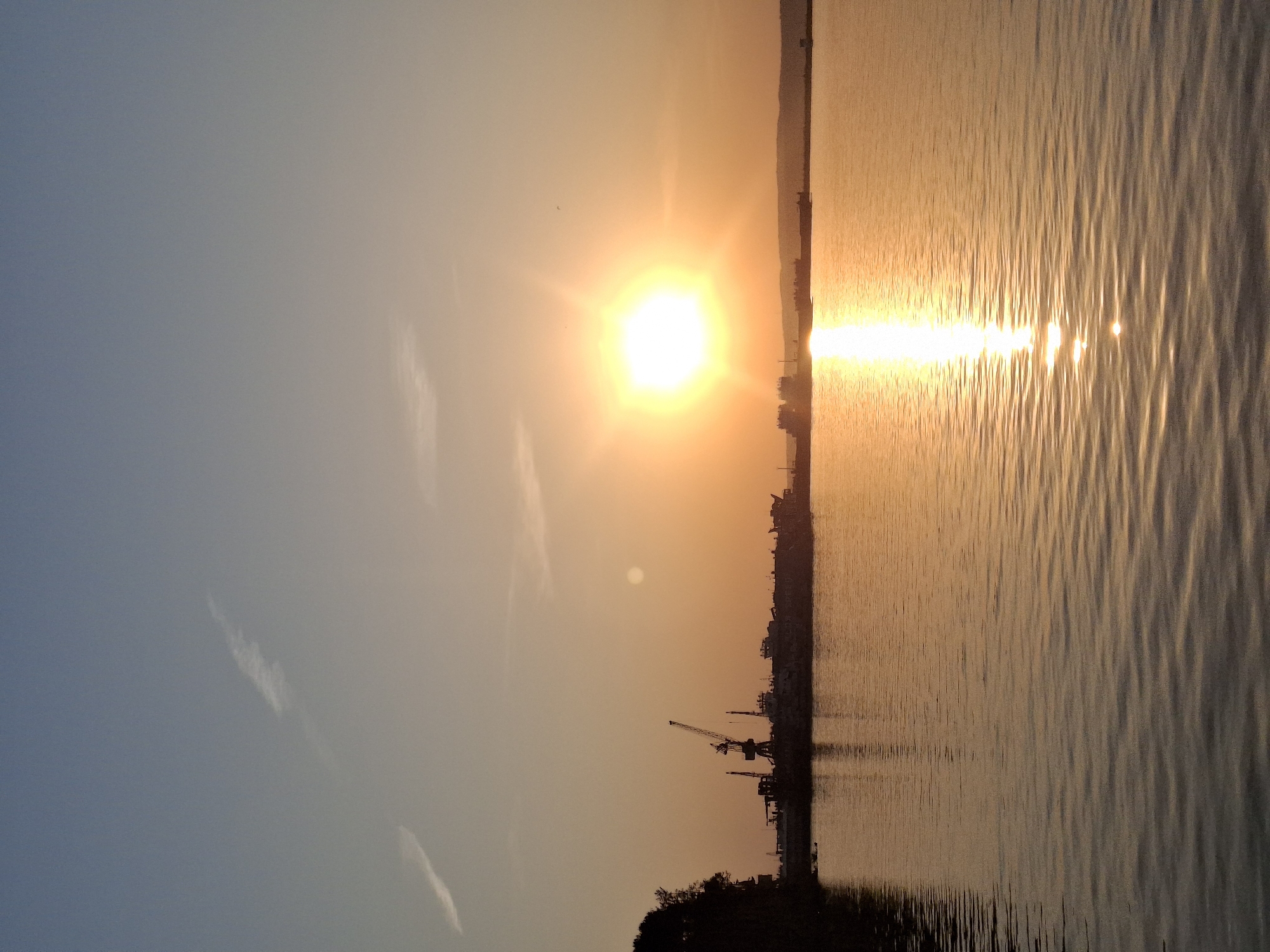 The image shows a sunset over water, likely a river or bay. The sun is a bright orb, reflecting intensely on the water's surface. Silhouetted against the sunset are land features including trees and what appears to be a skyline with industrial structures, possibly cranes. The sky ranges from a blueish hue to an orange-yellow gradient as it approaches the horizon. Given the city location as Chaikovsky, Russia, it is possible that this is the Kama River.