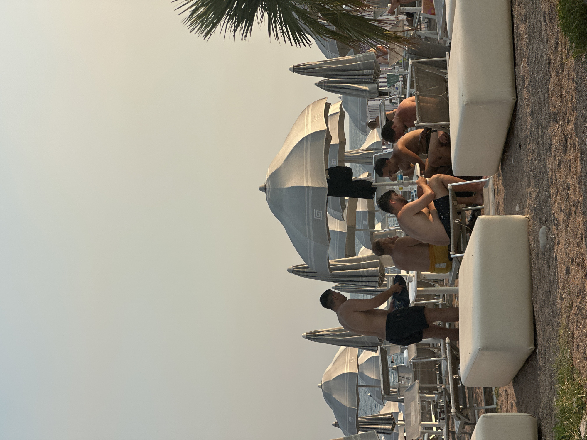 The image depicts a beach scene in Boreti, Montenegro, bathed in the soft light of late afternoon or early evening. The foreground is dominated by several large, white, rectangular ottomans placed on a pebble-covered ground. Beyond these, beach chairs and tables are arranged in rows, some occupied by people. Numerous closed beach umbrellas, primarily grey and white, stand in clusters, suggesting a beach resort setting.
In the midground, a group of four men are seated around a table, seemingly engaged in conversation or a game. One man, wearing black shorts, is standing and appears to be attending to something on the table. To his right, another man is wearing yellow shorts. The sea is visible in the background, calm and extending towards the horizon under a hazy sky. A palm frond, illuminated by the sun, is visible in the upper right corner, adding a tropical element to the scene. The overall atmosphere is relaxed, characteristic of a beach leisure area.