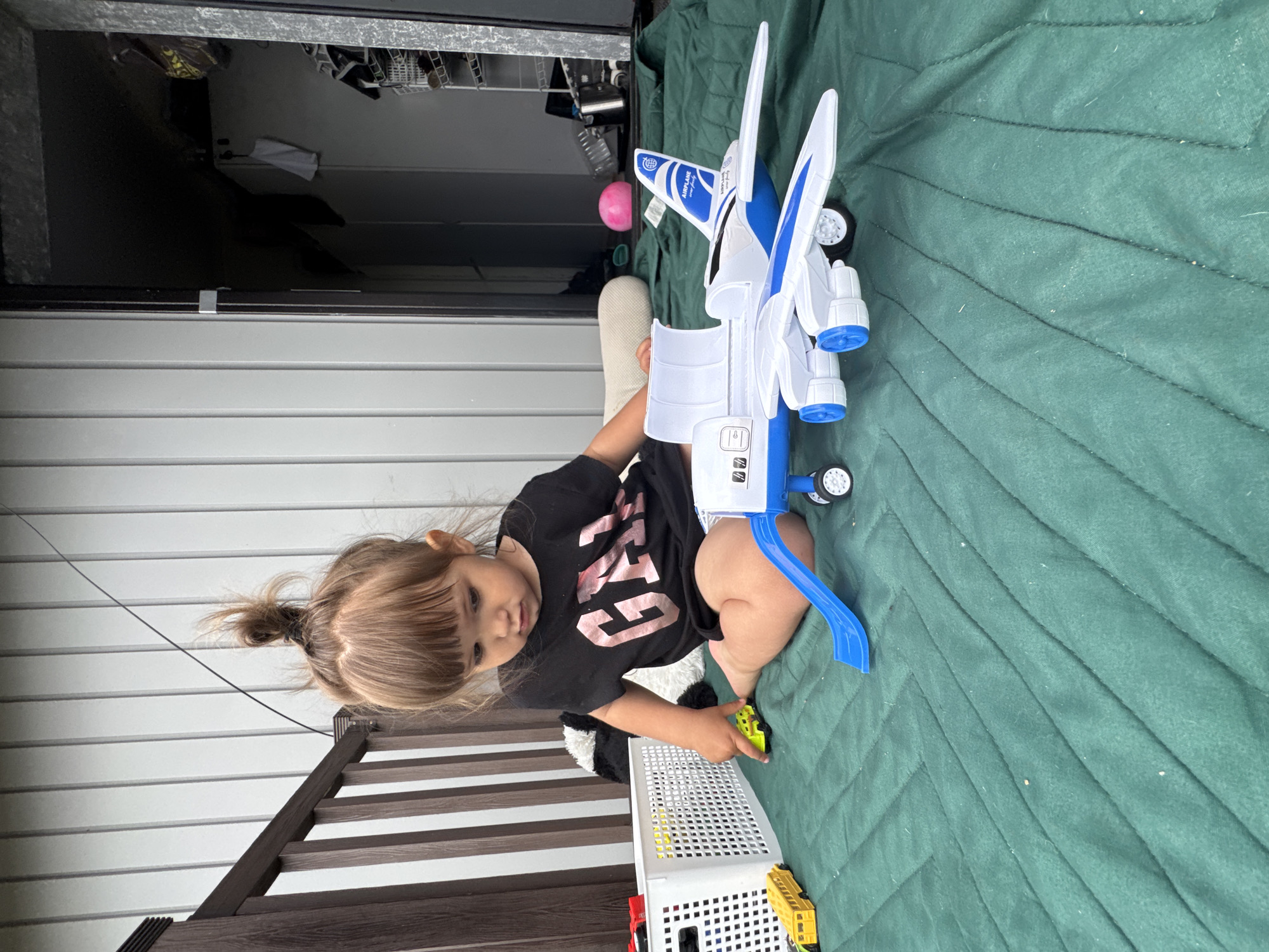 A young Caucasian child with blonde hair in a messy bun is sitting on a quilted green mat outdoors, playing with toys. The child is wearing a black t-shirt with "GAP" printed in pink letters. To the child's right is a white and blue toy airplane with open doors and a blue slide attached to its side. In front of the child and to the left is a white plastic bin filled with toys and a yellow toy school bus. Further left, another yellow toy vehicle is visible. The background consists of a light-colored, slatted wall, a wooden railing, and an open doorway leading to an interior space with shelves and various items. The scene appears to be on a balcony or patio. The location is identified as Kempelevo, Russia.