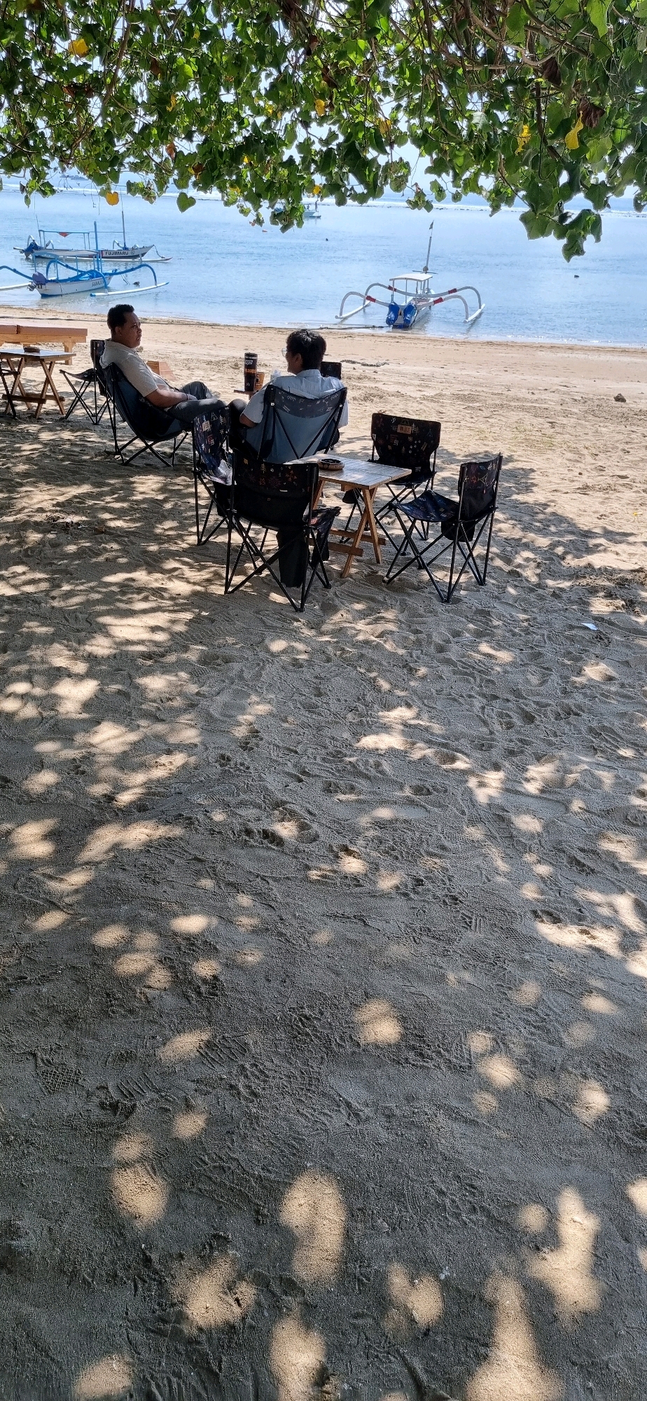 The image depicts a tranquil beach scene in Denpasar, Indonesia, under the dappled shade of trees. Two individuals are seated in folding chairs at separate small tables on the sand. The person on the left, a man wearing a light-colored collared shirt and dark pants, faces towards the sea and is looking to his right. The person on the right, wearing a light blue collared shirt and dark pants, is seated in a similar chair, facing away from the camera towards the sea, with their back partially to the viewer.
In the foreground, the sandy beach is covered in footprints and shadows cast by the overhead foliage. Scattered on the sand are several folding chairs and small portable tables. Two of these tables are positioned between the two seated individuals, with one table holding two drink containers.
Beyond the beach, the calm, blue ocean stretches to the horizon, with several traditional Balinese fishing boats, known as jukungs, visible in the water. The boats are characterized by their outriggers, which help maintain stability. One boat in the mid-ground is white with blue accents, while another further back is predominantly blue with white accents. The water appears relatively shallow near the shore. The sky is clear and bright, indicating a sunny day. The overall atmosphere is one of relaxation and leisure.