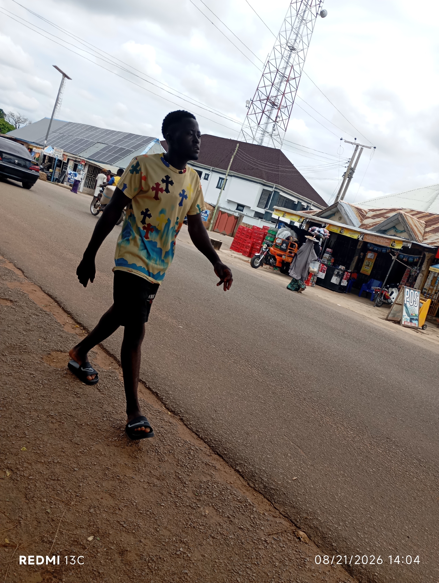 In the city of Shendam, Nigeria, a young Black male with dark skin, short black hair, and wearing a light-colored t-shirt with a cross pattern and black shorts walks on a paved road. He is also wearing black slide sandals. In the background, there are buildings, a cellular tower, solar panels, parked motorcycles, and a person with a gray covering on their head walking with items on their head. The environment appears to be a commercial area with shops. The image was captured on August 21, 2026, at 2:04 PM.