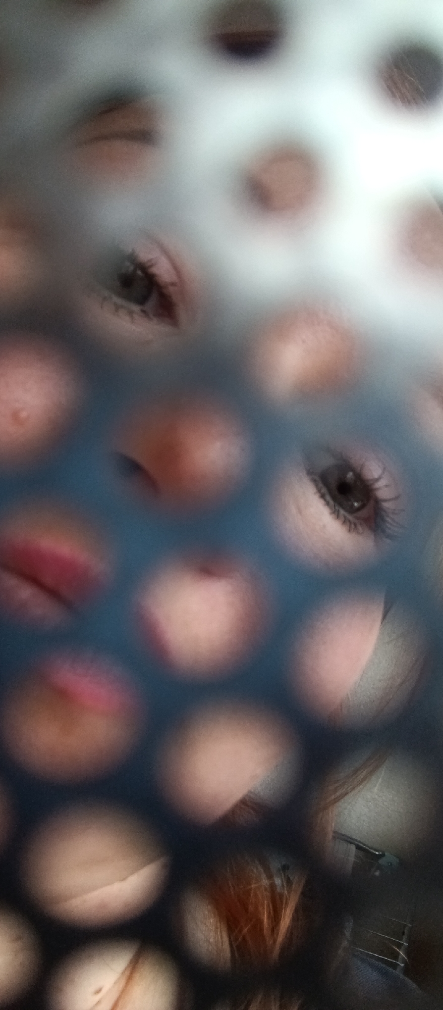 A close-up photograph captures a person's face viewed through a perforated screen or object, creating a fragmented, mosaic-like effect. The face is divided into numerous circular segments, revealing portions of the eyes, nose, and lips. Both eyes are visible through the circular openings, with notable dark eyelashes and irises. The bridge of the nose and reddish lips are also discernable through other perforations. The skin tone appears fair to medium. In the lower right corner, strands of reddish-brown hair are visible. The foreground object, which is dark-colored (possibly blue or black) and perforated with circular holes, is the primary visual element, obscuring and framing the facial features. The background is indistinct and blurry, suggesting an indoor setting. No specific activity or event is taking place beyond the photographic capture of the face. The time of day and weather are undeterminable, and there is no visible text.