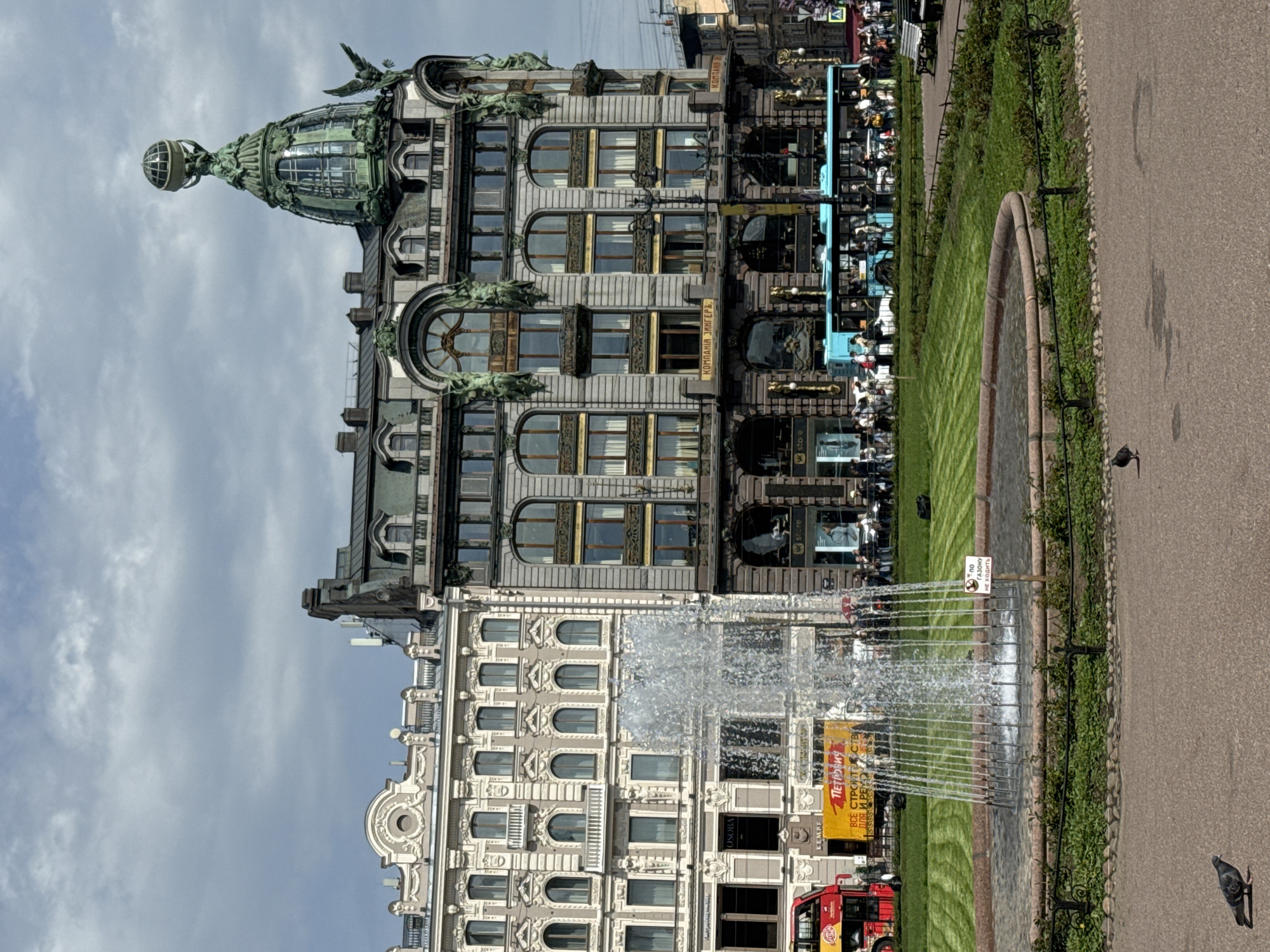The image features the Singer House, located on Nevsky Prospect in Saint Petersburg, Russia. The architecture is notable for its Art Nouveau style and its glass dome topped with a globe. The building appears to have a dark gray facade with ornate detailing. Below, a fountain is present in a landscaped area with grass and a paved walkway. A double-decker bus is visible near the fountain. The fountain is a modern, linear design. A sign is visible in Russian. The sky is partly cloudy. Pedestrians are visible in the background.