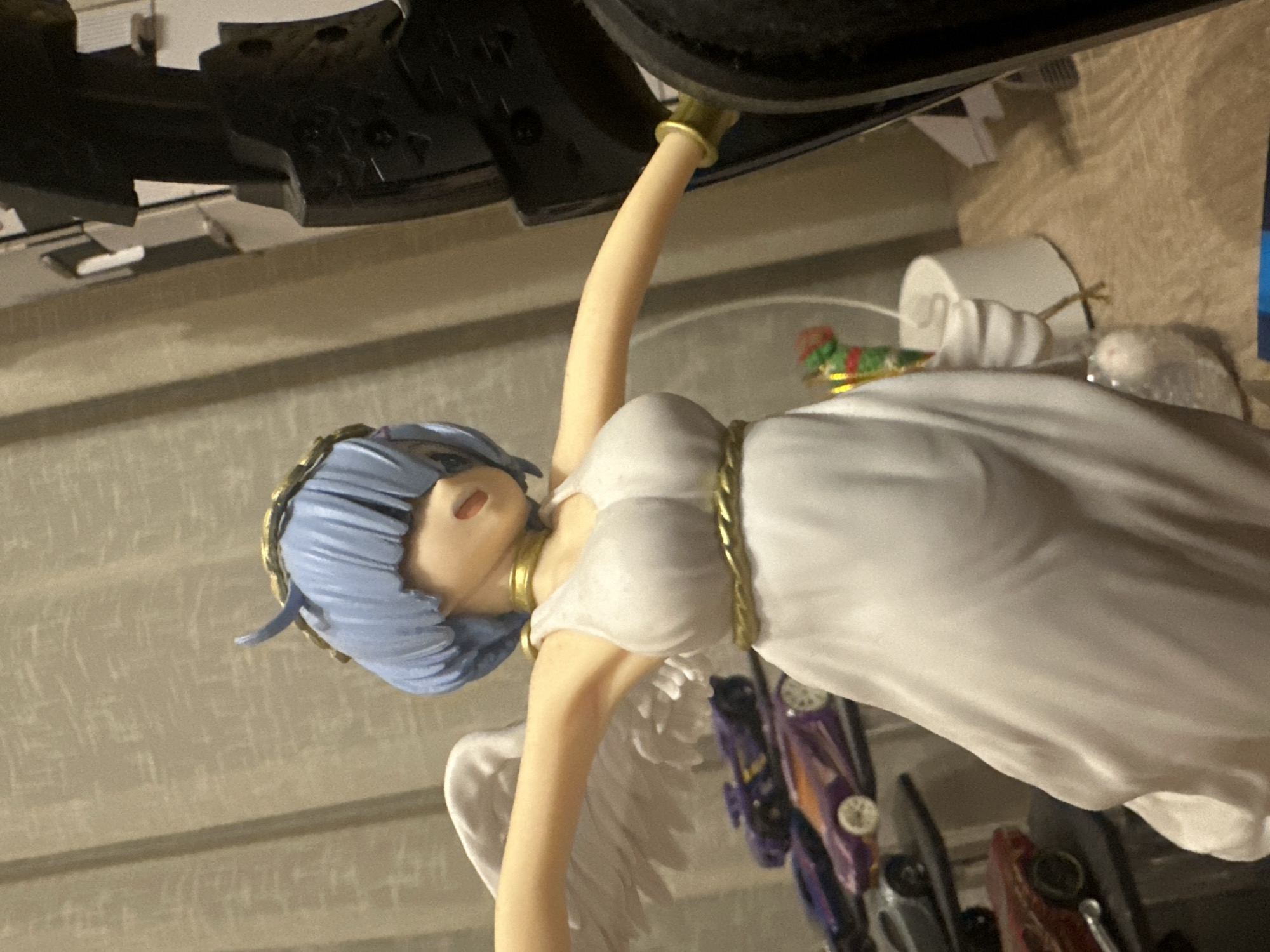 An angel figurine with blue hair, a golden halo, and white wings stands in front of a beige curtain. The figurine is wearing a white dress with a golden rope belt. To the left of the figurine are several toy cars on shelves. To the right, a portion of a black, large object is visible. The scene is indoors, with a textured surface in the background.