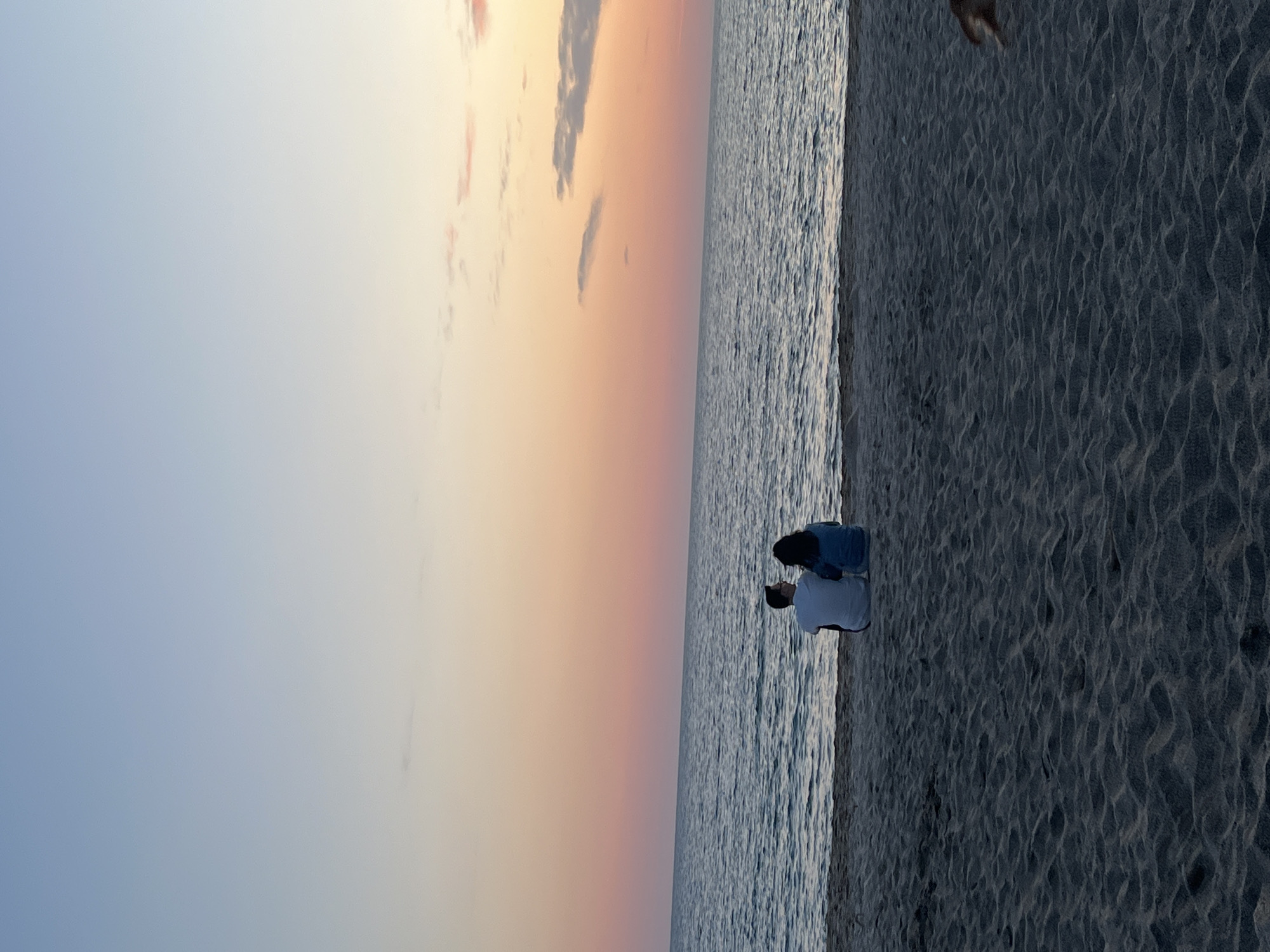 The image depicts a tranquil beach scene in Alanya, Turkey, likely during sunset or sunrise. The expansive sky dominates the upper portion of the frame, showcasing a beautiful gradient of colors transitioning from a soft, light blue at the top to warm oranges, pinks, and faint yellows closer to the horizon. A few wispy clouds are visible, adding texture to the colorful sky.
Below the horizon, the calm ocean stretches out, its surface exhibiting gentle ripples that reflect the ambient light. On the sandy beach, two individuals are positioned near the water's edge. They appear to be seated or kneeling, with their backs partially facing the viewer, as they gaze out towards the sea and the glowing horizon. One person wears a light-colored top, possibly white or light blue, while the other is dressed in a darker blue or denim top. The sand in the foreground and extending along the shoreline displays intricate, wavy patterns, characteristic of a beach where the tide has recently receded. No visible text is present in the image. The overall mood is serene and contemplative, capturing a peaceful moment by the sea.