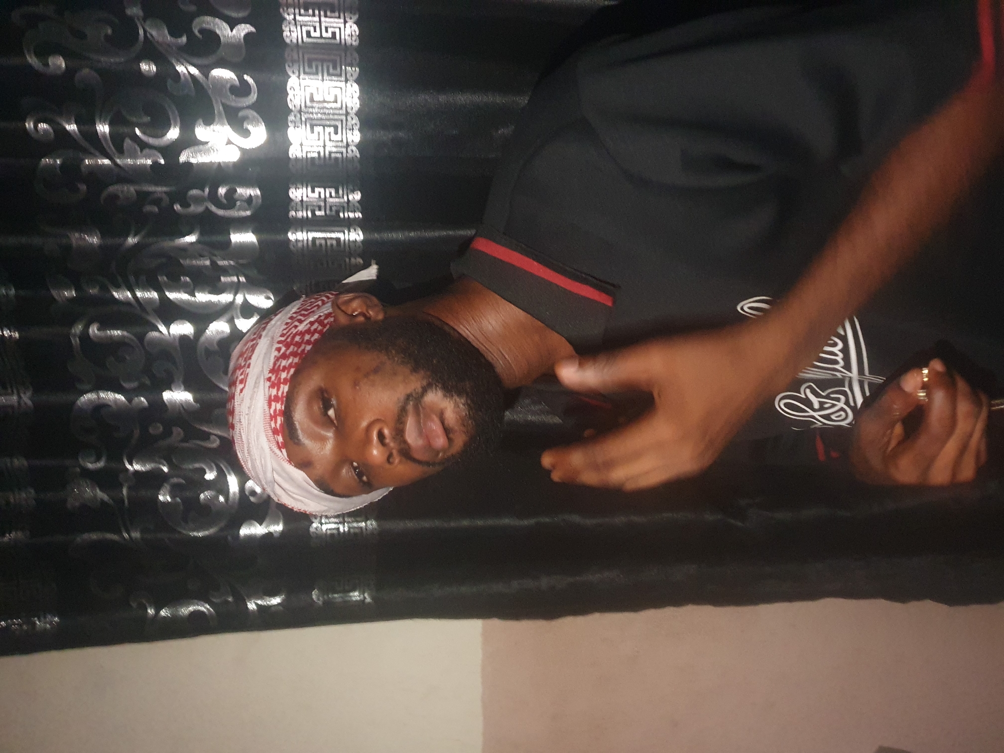 A medium shot depicts a man with dark skin, a black beard, and a red and white patterned head covering. He wears a black shirt with a red collar trim and a white graphic on the chest. He is facing the camera with a slight tilt to his left, and his right hand is raised. Behind him is a black, patterned fabric, suggesting a backdrop. The lower portion of the image shows a transition to a neutral-toned surface, possibly a wall or floor. The location, Lokoja, Nigeria, is not visually apparent and can only be inferred if it is known that the photo was taken there.