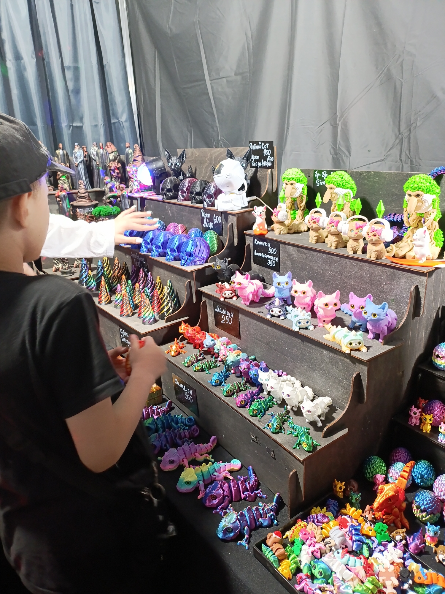 The image shows a young person with dark hair, wearing a black t-shirt and a black cap, looking at a display of colorful toys in a store. The toys are arranged on tiered shelves and in trays. The items include many small, brightly colored plastic figures, such as unicorns, cats, chameleons, and possibly mythical creatures. Some larger items, like skulls and figures with green hair, are also visible. A white-clad person's hand is reaching out towards some blue, iridescent skull figures. The background consists of gray curtains. The scene appears to be a vendor stall at a market or convention, given the display of merchandise. The location is identified as a store in Lagunovo, Russia.