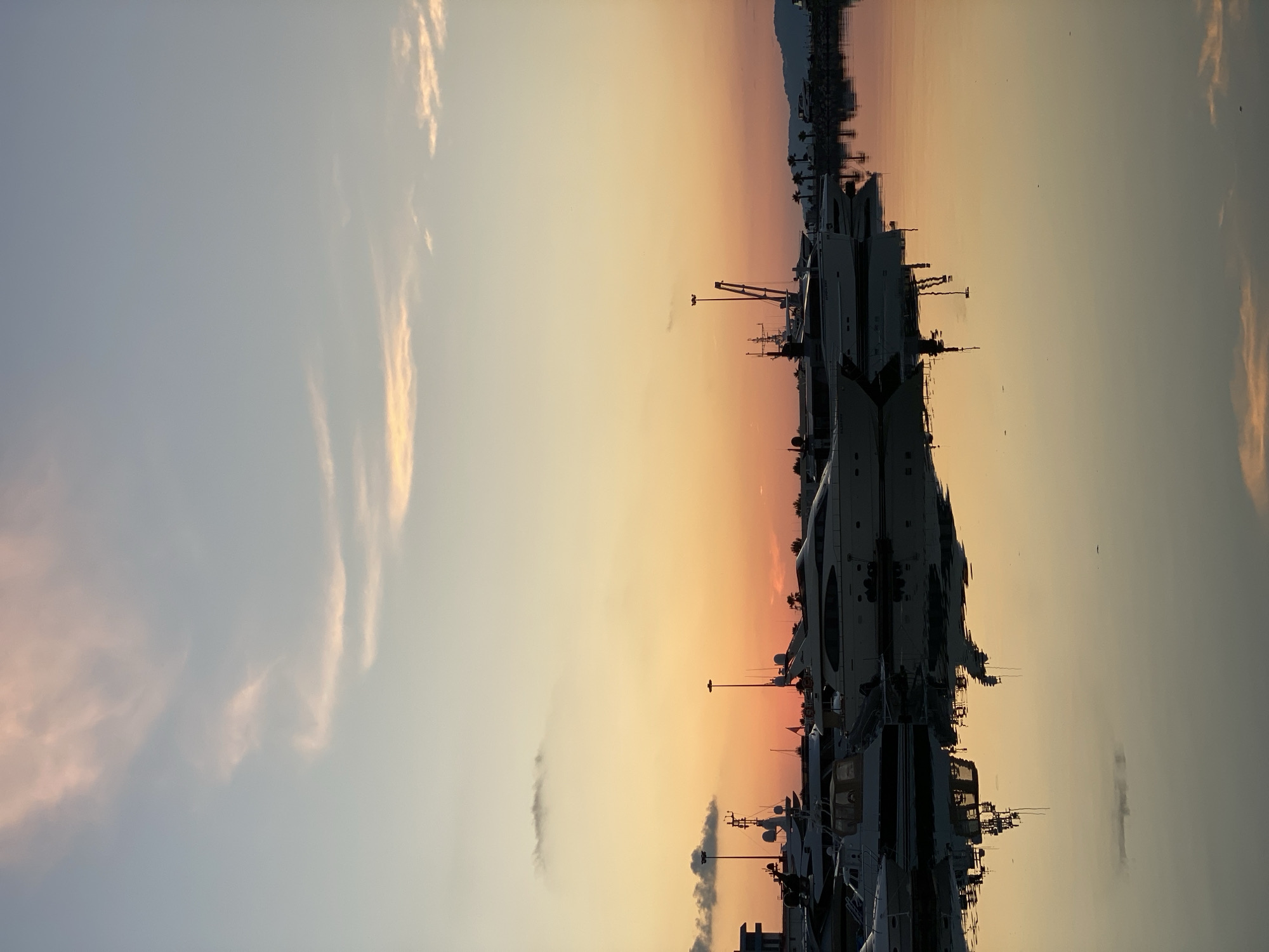 A serene sunset scene unfolds at the Sochi, Russia, marina. The sky transitions from a pale blue at the top to a warm gradient of orange and pink near the horizon, dotted with wispy clouds. Several yachts, varying in size and design, are moored in the calm water, their reflections shimmering on the surface. The silhouettes of palm trees and distant mountains are visible against the colorful sky. The overall atmosphere is peaceful and tranquil, typical of a harbor at dusk.