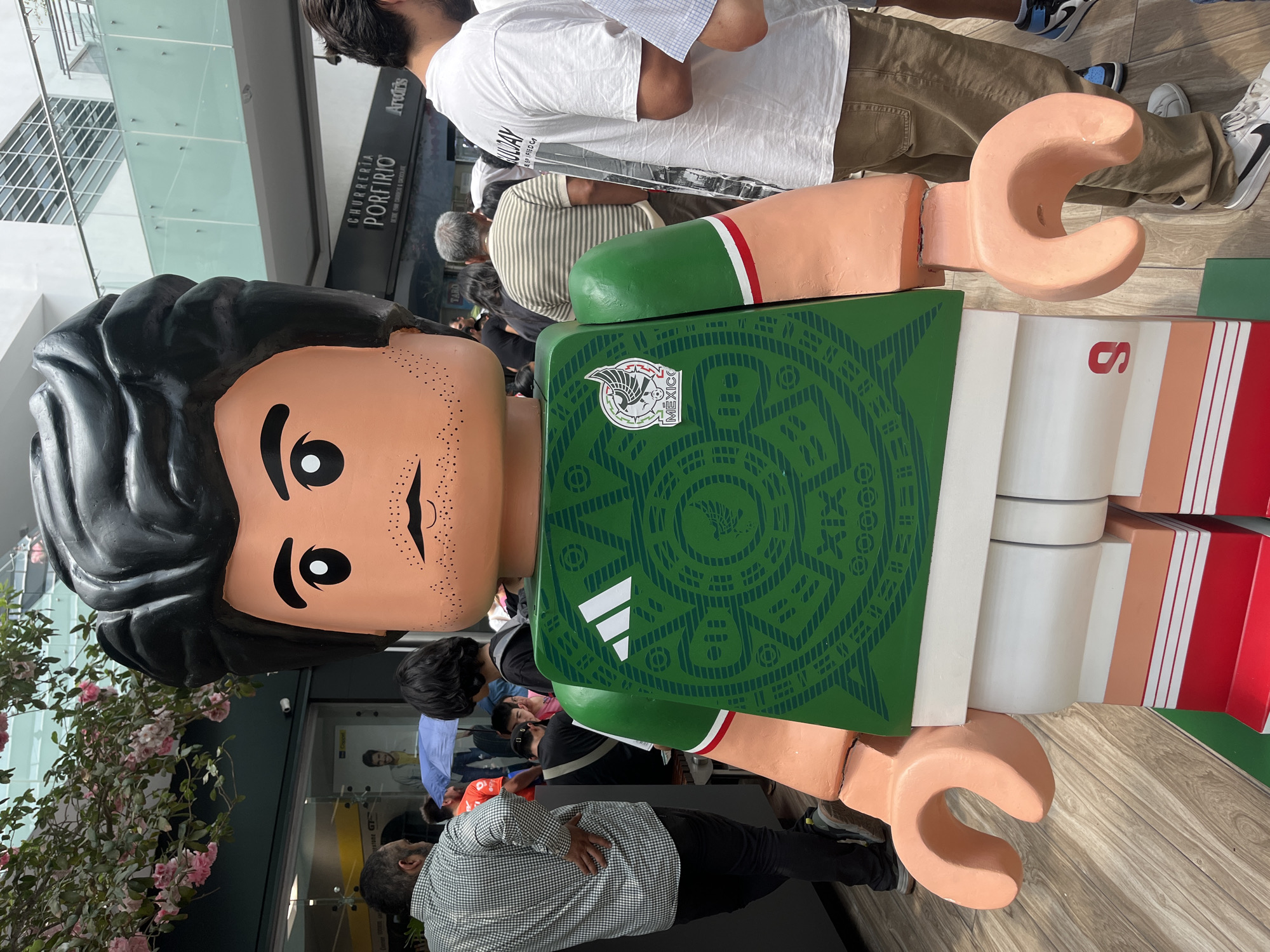 The image captures a life-sized LEGO figure of a soccer player, specifically designed with elements representing the Mexican national team. The figure stands outdoors, with a crowd of people visible in the background, suggesting a public exhibition or event.
The LEGO figure has a classic blocky design, with a tan face, simple black eyes, and a faint mustache. Its hair is dark, styled in a somewhat spiky manner. The torso of the figure is painted with a green jersey bearing the Adidas logo and the emblem of the Mexican national team, adorned with a detailed pattern. The figure has blocky, hook-shaped hands in a tan color. The lower part of the figure, presumed to be the legs, features white and red stripes, hinting at the colors of the Mexican flag.
The backdrop includes parts of a building with glass panels and a sign that reads "CHURRERIA PORFIRIO DESDE 1909 CHURROS & CHOCOLATE," and "PORTIRO." The crowd seems to be observing or interacting with the LEGO figure, with several people partially visible behind and around it. Some are taking pictures, while others are simply watching. There are flowering bushes nearby.
The scene is well-lit, suggesting it is daytime. The location appears to be in the city of Unknown, Unknown based on the OCR of the image.