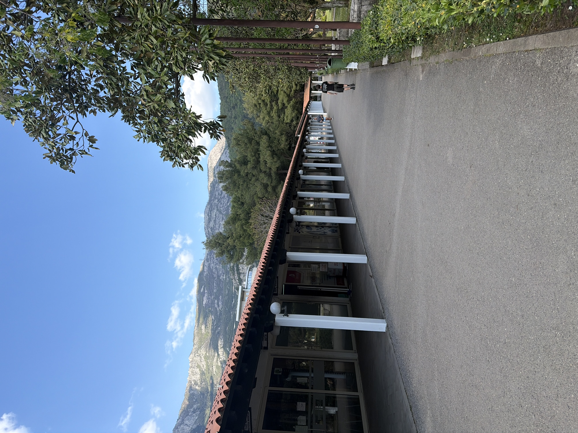 The image depicts an outdoor pedestrian walkway in Budva, Montenegro, under a bright blue sky. On the left, a vibrant green tree canopy partially frames the scene. A wide asphalt path extends from the foreground into the distance, running parallel to a long, single-story building on its right. This building features a distinctive terracotta tiled roof and a series of white pillars that support an overhang, creating a sheltered arcade. Its facade comprises multiple glass storefronts, suggesting commercial establishments.
In the mid-ground, a person wearing a dark t-shirt and shorts walks away from the camera along the paved path. Another individual is partially visible further down near the building's entrance. On the opposite side of the path from the building, a row of vertical wooden or metal poles forms a trellis-like structure, beyond which are green bushes and a small green trash can.
The background is dominated by majestic, rugged mountains, some with exposed rock faces and others covered in lush greenery, stretching across the horizon under a clear blue sky dotted with a few scattered white clouds. The weather is sunny and bright, indicating daytime. Text partially visible on one of the building's glass doors includes "ENERGETSKA AGENCIJ...", suggesting an energy agency or similar business. Other signs, while indistinct, imply commercial activity. The overall scene conveys a calm, everyday atmosphere in a scenic, mountainous, and potentially resort-like setting.