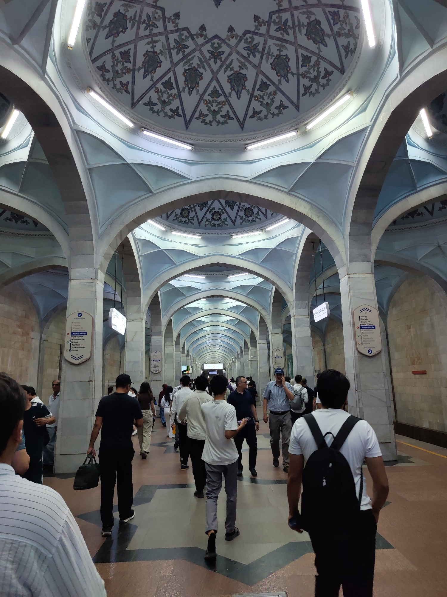 The image depicts a bustling scene within a richly decorated metro station in Tashkent, Uzbekistan. The architecture is characterized by a series of grand arches and a domed ceiling adorned with intricate patterns. The station features polished marble columns and pale blue vaulted ceilings, illuminated by long fluorescent lights. A long corridor stretches into the distance, lined with more arches, creating a sense of depth.
Numerous people are walking in various directions along the platform, which appears to be made of tile and stone. They are dressed in casual, modern clothing. Several individuals are carrying bags or backpacks. Informational signs with text in Latin and Cyrillic scripts are mounted on the columns, indicating station names and directions. The overall atmosphere is one of movement and activity within an ornate public space. The style of the station strongly suggests it is part of the Tashkent Metro system, known for its elaborate Soviet-era designs.