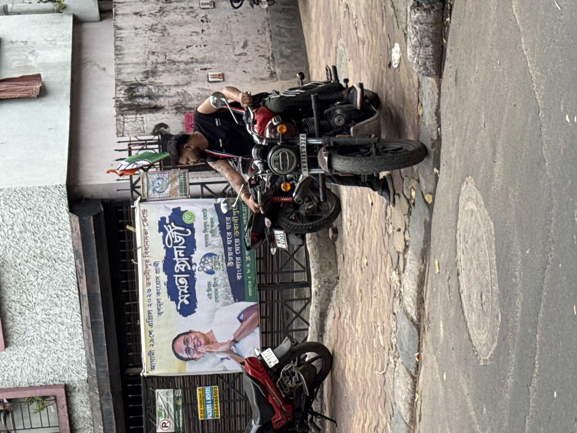 The image captures a street scene in Kolkata, India, featuring a young man and two motorcycles, with a prominent political banner in the background.
A man, wearing a black t-shirt with red and white stripes and dark trousers, stands on a tiled sidewalk next to a classic-style motorcycle. He appears to be interacting with the motorcycle's front section, possibly adjusting something, with his tattooed right arm visible. The motorcycle is dark-colored with maroon accents and features the license plate "WB 01AS 8324".
Behind the man, a large political banner is displayed on a building or gate. The banner features an image of Mamata Banerjee, a prominent Indian politician, smiling and gesturing. The Bengali text on the banner identifies her as the "Trinamool Congress candidate for Bhabanipur constituency on April 29, 2026." It includes a call to action: "Press the button on this symbol (Trinamool) and make Trinamool victorious again with a huge margin," and mentions "Development in every house, Bengal's daughter in Bhabanipur, Campaign: 82 Block Rupom Congre...".
Partially visible to the left is another motorcycle, red and black, with the license plate "WB 19 C-7376". Below the political banner, a small sign indicates "NO PARKING," along with "PACKERS & MOVERS" and a contact number "9564486503." Other numbers, "8100002772" and "8100004098," are visible on a white box near the gate.
The scene is set on a patterned tiled sidewalk that borders an asphalt road. Buildings with light grey, textured walls form the backdrop. The clear, bright lighting suggests it is daytime.