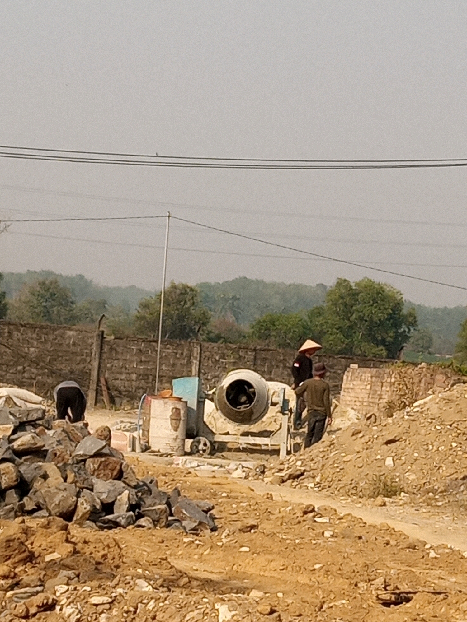 The image depicts an outdoor construction site in Teluk Dalem Ilir, Indonesia. Three individuals are present. One person, wearing dark pants and a grey shirt, is bent over near a pile of large stones in the foreground. To the right of this person, a cement mixer is visible. Two other individuals are near the cement mixer; one, wearing a conical hat and dark clothing, stands beside it, while another person in dark clothing stands further to the right. A low brick wall runs along the background, with trees and foliage beyond it. Electrical wires are strung across the upper portion of the image. The ground is uneven, with piles of dirt and stones. The overall scene suggests ongoing construction work.