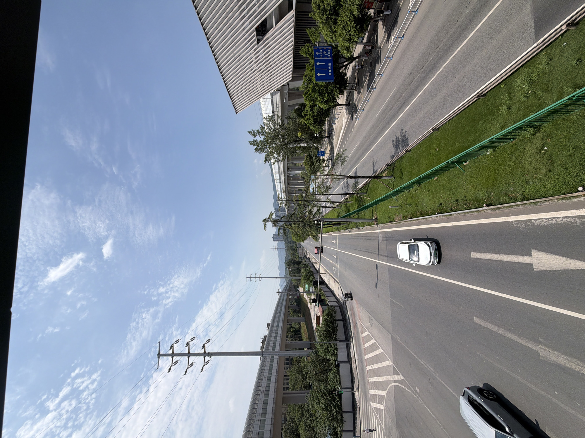 The image captures a daytime, elevated perspective of a multi-lane road in Tiaodeng, China. The road is flanked by green grassy areas and metal fencing. On the left, a modern elevated train or monorail structure with a covered walkway runs parallel to the road. Large electrical pylons with power lines are visible to the left of the road, with a white car and a grey car on the road. A pedestrian crossing is marked on the road. On the right side of the road, a large, contemporary building with a distinctive sloped, metallic roof is present. Traffic signs, including directional arrows and Chinese characters, are posted on the right side of the road. Several trees line the sides of the road and the area around the building. Two vehicles are seen traveling on the road: a white car in the right lane and a red bus further down the road. The sky is blue with scattered white clouds.