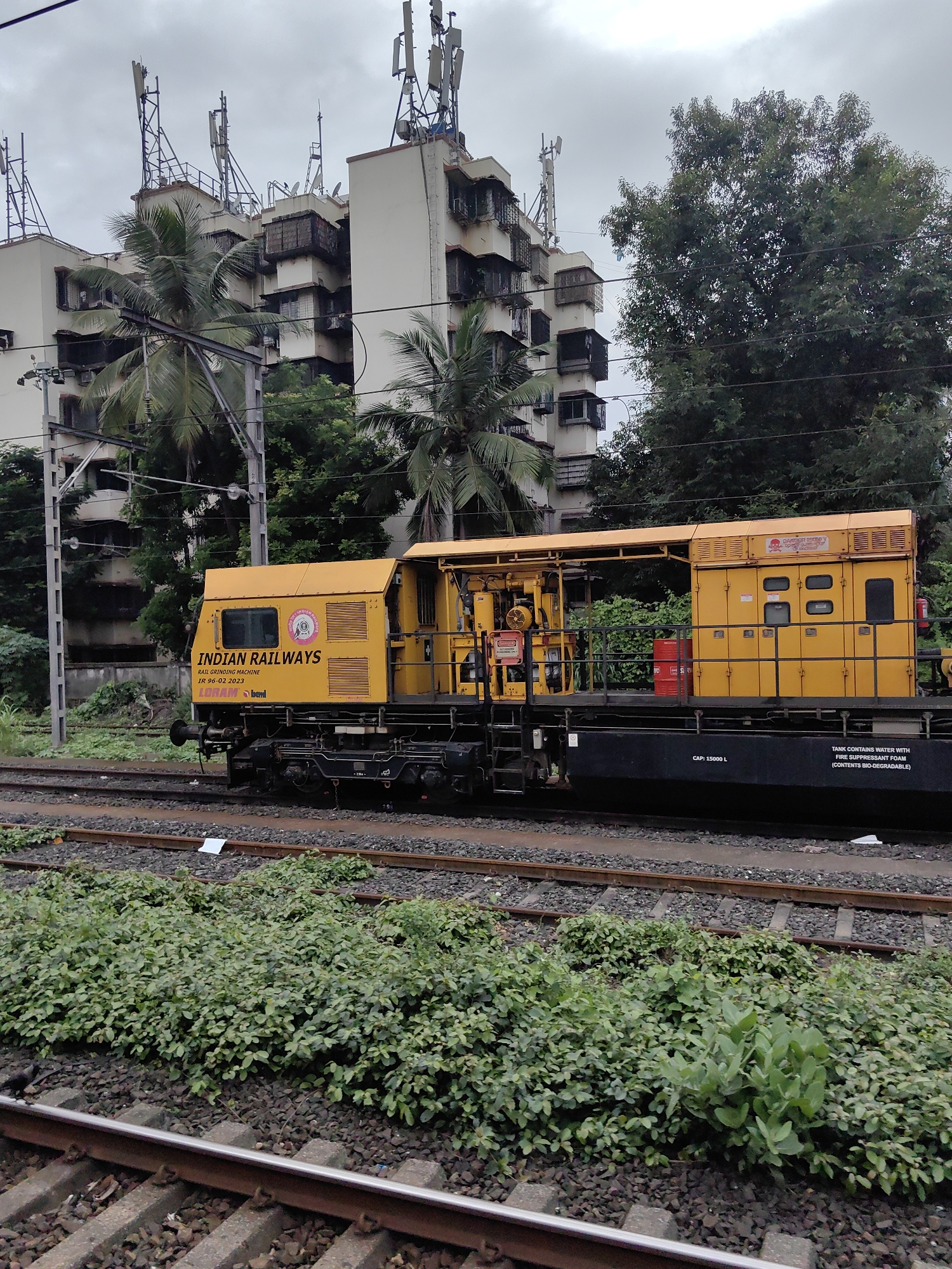 A yellow Indian Railways rail grinding machine is parked on train tracks in a dense urban environment. The machine has "INDIAN RAILWAYS" prominently displayed in black lettering. Behind the train, a multi-story apartment building is visible, characterized by its light-colored facade and numerous windows. Several cellular antennas are mounted on the roof of the building. Palm trees and other lush green foliage surround the building and railway tracks, indicating a tropical or subtropical climate. The sky is overcast with grey clouds. The tracks are surrounded by ballast and vegetation. A black car, possibly a maintenance vehicle, is partially visible behind the yellow machine. The overall scene suggests an industrial or transportation hub within a residential area.