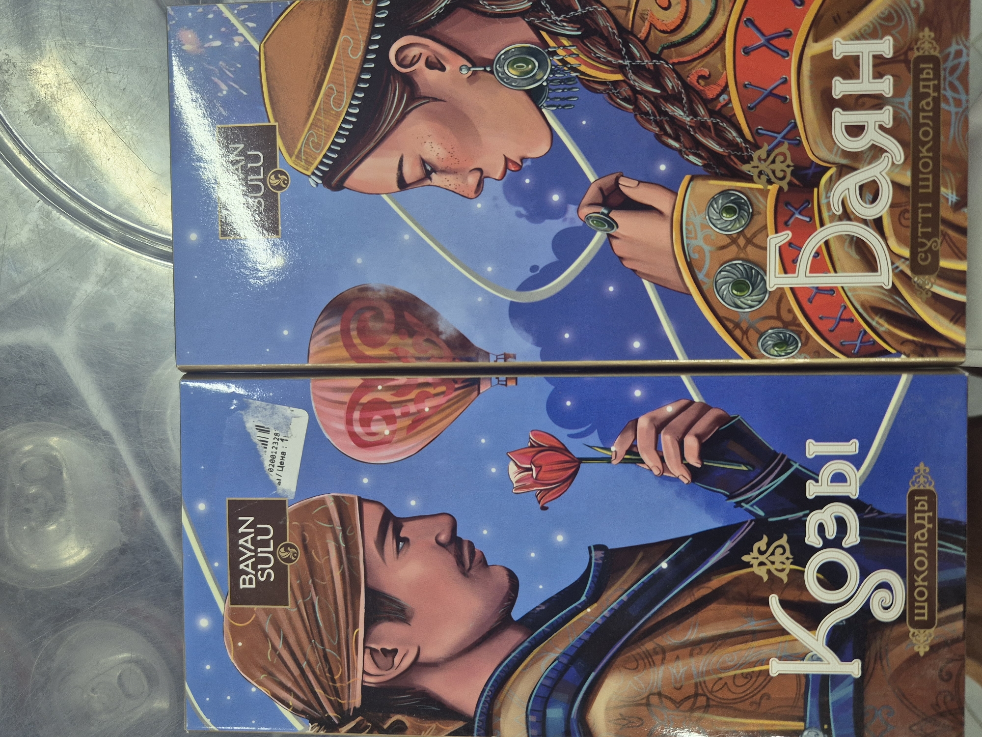 Two chocolate boxes are displayed side-by-side. Both boxes feature artistic illustrations of a man and a woman in traditional Kazakh attire against a starry night sky with hot air balloons. The left box has the brand "BAYAN SULU" at the top and the word "Қозы" in large, decorative font, with "ШОКОЛАДЫ" below. The right box also has "BAYAN SULU" at the top and the word "Баян" in large, decorative font, with "сұтті шоколады" below. The man on the left holds a red tulip, while the woman on the right looks away. Both figures are depicted in profile. The background on both boxes is a deep blue with white stars and wisps of clouds. The boxes are placed on a metallic surface, possibly a shelf or table, with visible circular patterns suggesting an industrial or storage setting. There are no visible people present in the scene, only the illustrated figures on the packaging. The overall impression is a product display. The city of Kokshetau, Kazakhstan is known to be the location of the Bayan Sulu confectionery factory.