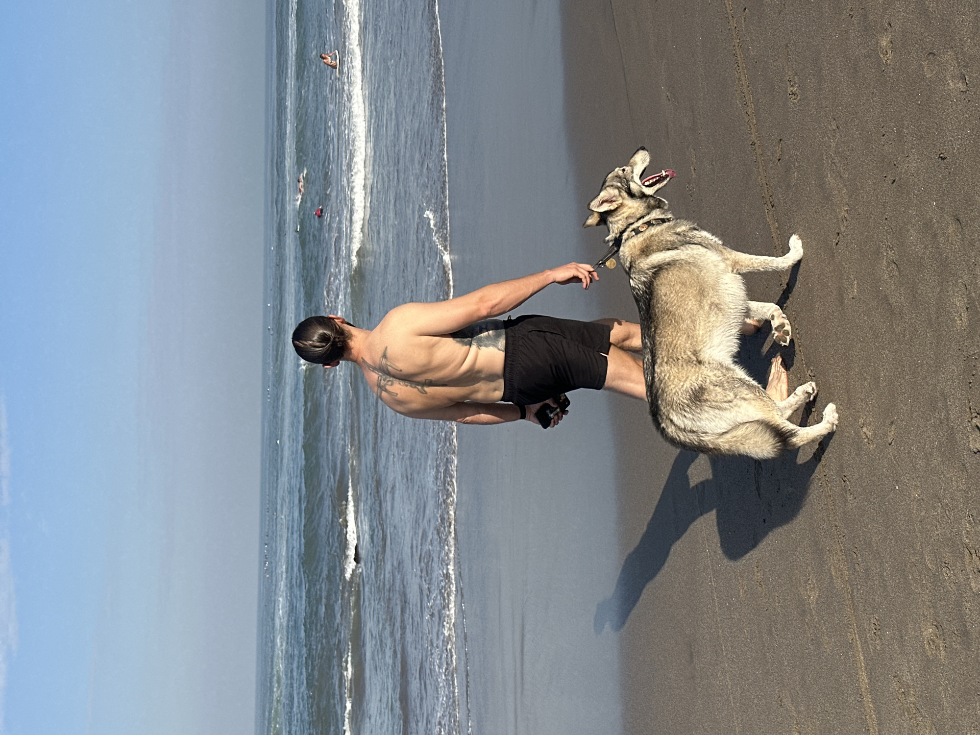 An adult male and a dog walk along a sandy beach shoreline in North Kuta, Indonesia. The shirtless male, observed from behind, has a top knot hairstyle and extensive tattoos covering his back and left arm. He wears black shorts, holds a smartphone in his right hand, and the leash for the dog in his left. A light brown and grey dog, consistent with a husky or wolf-dog breed, walks on a leash to the man's left, slightly ahead. The dog's mouth is open with its tongue extended, and it wears a collar with a visible tag. The immediate foreground consists of wet, dark sand exhibiting footprints and a distinct long shadow extending from the man and dog towards the left. The adjacent ocean features breaking waves with white foam along the shore and deeper blue water extending to the horizon. Multiple individuals are visible further offshore in the ocean, positioned on surfboards, suggesting surfing activity. The sky above is clear light blue, with a subtle haze near the ocean horizon.