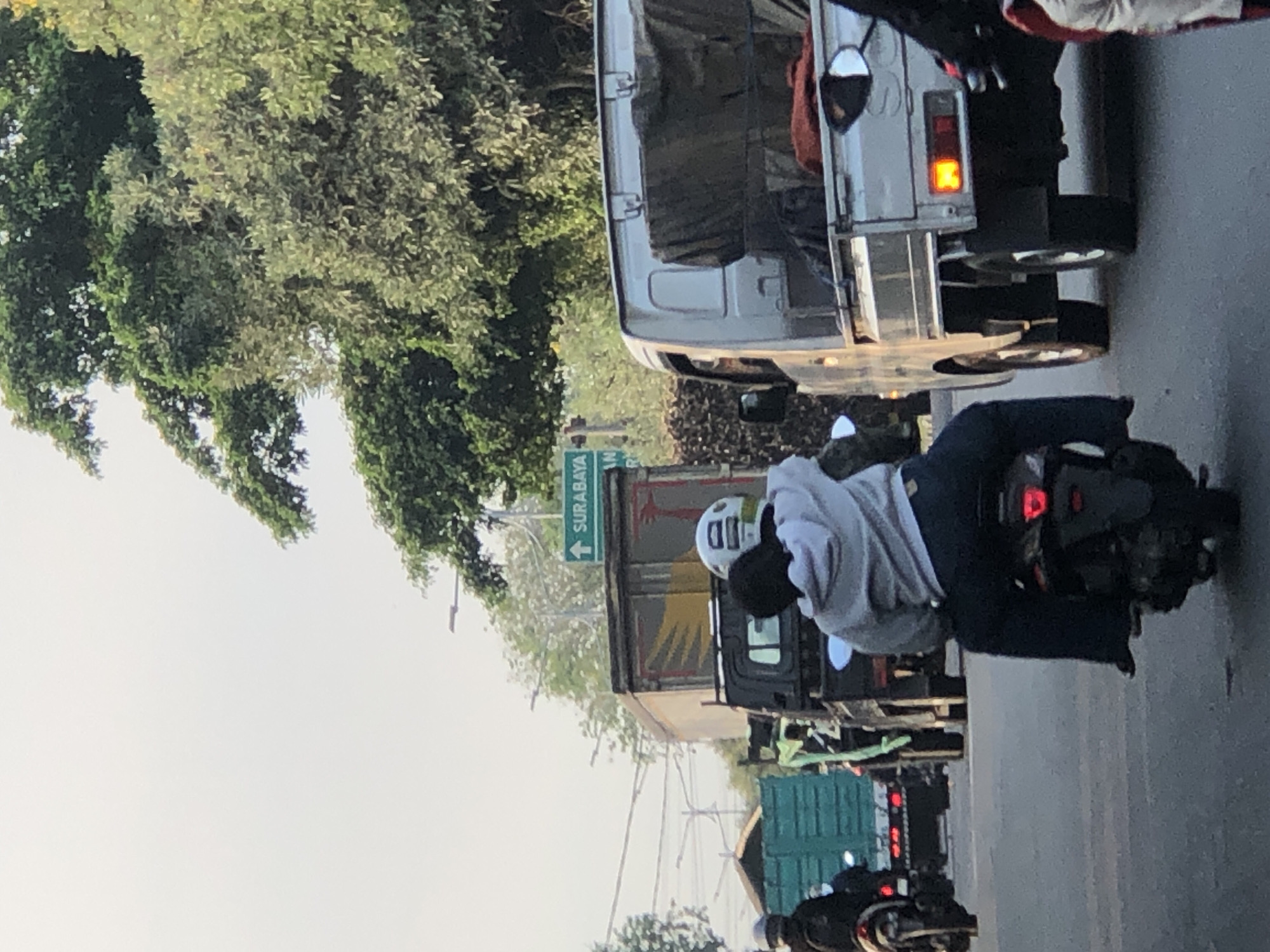 The image depicts a traffic scene on a paved road, likely in or near Kludan, Indonesia. The sky is overcast, suggesting it's either early morning or late afternoon. The road is congested with several vehicles, including motorcycles, trucks, and a large green truck in the background.
In the foreground, two individuals are riding a motorcycle. The person in the back is wearing a gray hoodie and dark pants, with their arms around the person in front. The rider in front is wearing a helmet. Another motorcycle is visible to the left, with a person wearing a dark helmet and jacket.
In the middle ground, two trucks are stopped in traffic. The truck on the left is a dark-colored truck with a yellow and red emblem on its side. The truck on the right is a lighter-colored truck with its bed covered by a tarp. A green truck is parked further back on the left.
A green sign with white lettering indicates "SURABAYA" with an arrow pointing upwards, suggesting this is a road leading to the city of Surabaya. Trees and vegetation line the sides of the road, indicating a somewhat suburban or semi-rural setting within the urban landscape. The overall impression is of a typical day in a busy Indonesian city, with traffic and everyday activities taking place.
