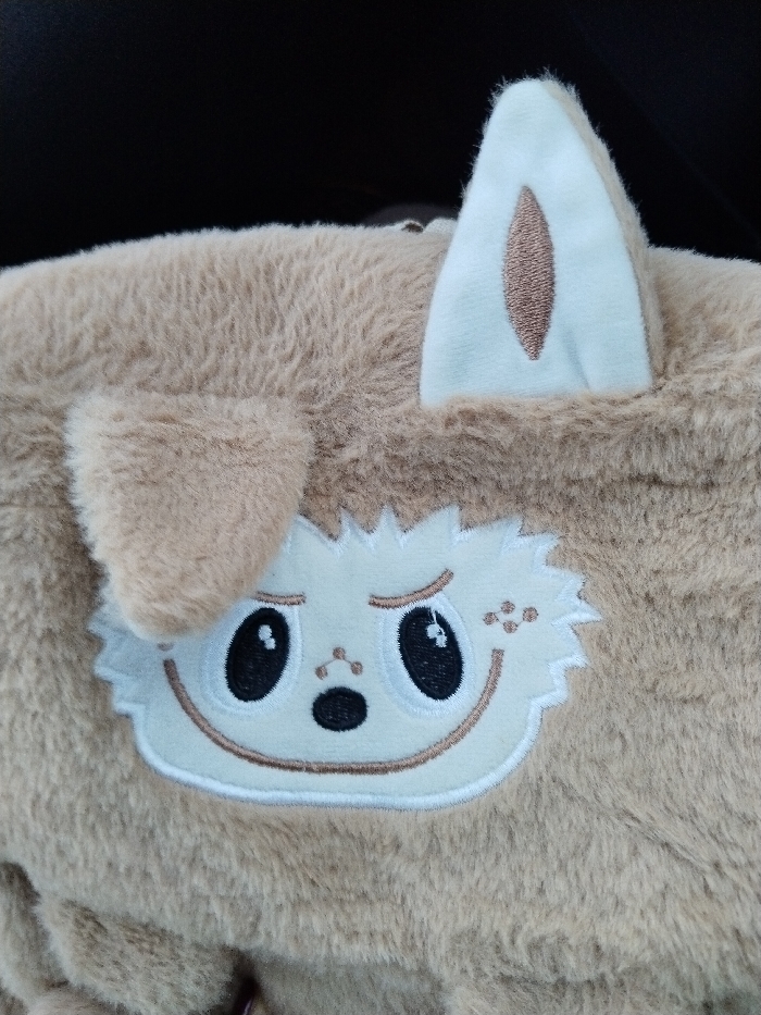 A light brown, furry plush toy is visible. It features an embroidered face with large black eyes, a small nose, and a simple mouth. On the upper left side of the face, there's a small, round, plush ear. On the upper right side, a white, triangular ear stands upright, with a brown stitched detail inside. The background is dark and blurry, indicating an out-of-focus setting.