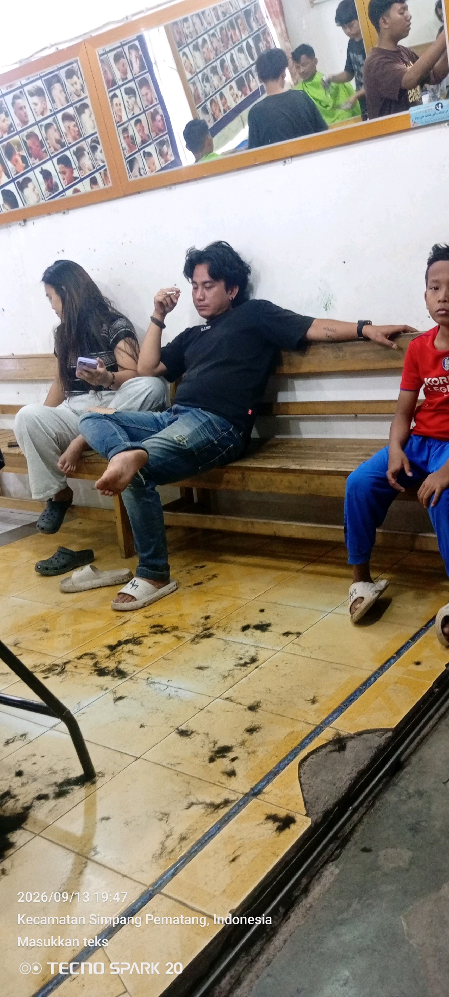 The image depicts the interior of what appears to be a barbershop in Simpang Pematang, Indonesia. The floor is tiled in a yellow and beige pattern and is scattered with dark clippings of hair, indicating recent haircutting activity. Three individuals are seated on a wooden bench. On the left, a woman with long dark hair is looking down at a mobile phone she is holding. Beside her, a man with dark, wavy hair is leaning back on the bench, smoking a cigarette and wearing a black t-shirt and blue jeans. To the right of the man, a young boy is seated, wearing a red t-shirt and blue pants. In the background, a mirror reflects a barber at work, indicated by a green cape on a person being cut, and a display of hairstyles on the wall. The date and time stamp indicates the photo was taken on 2026/09/13 at 19:47.